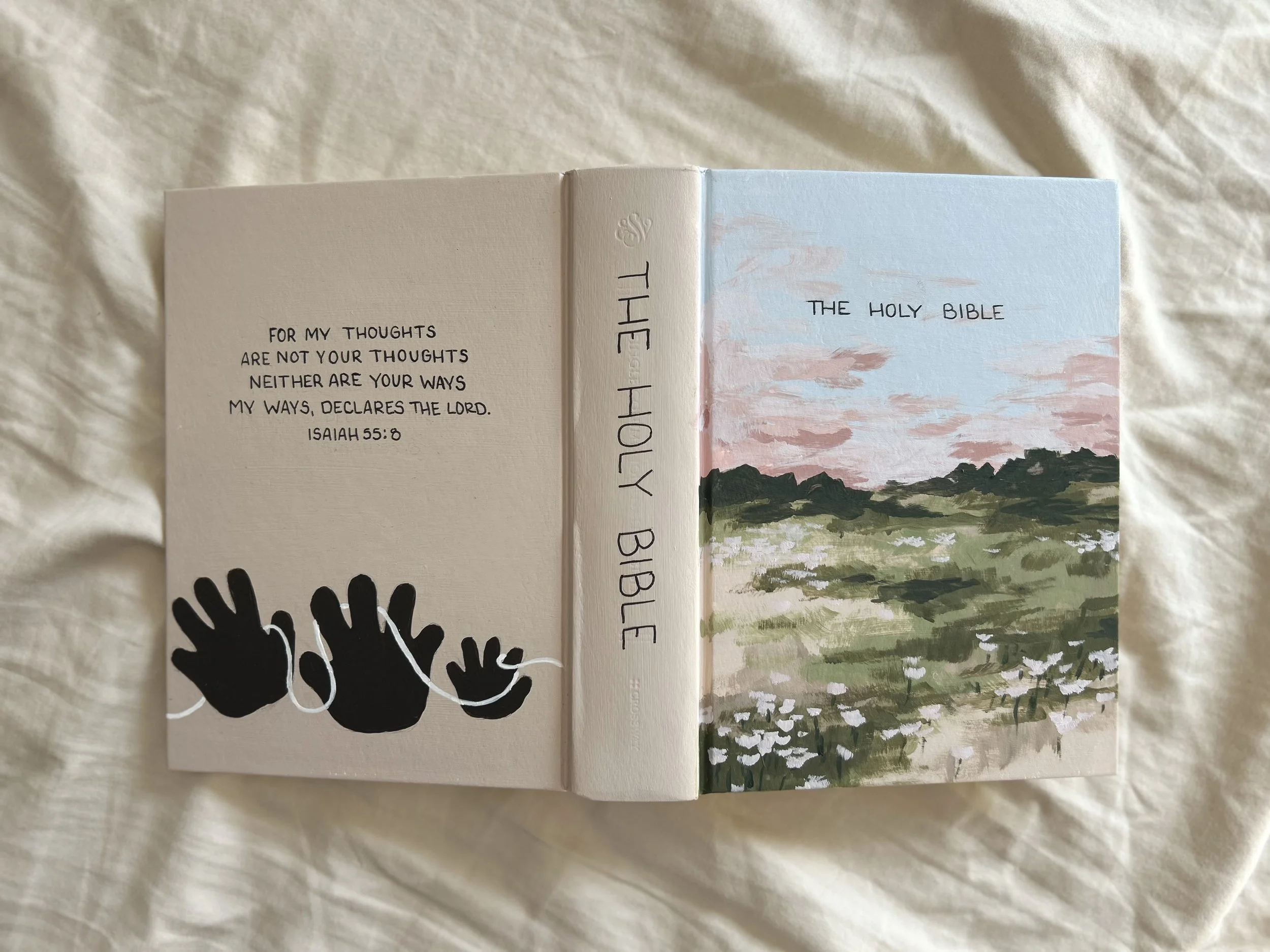 Open Bible with verse Isaiah 55:8 on the left page, and a landscape painting with trees and clouds on the right page, with black and white hand illustrations in the bottom left corner.