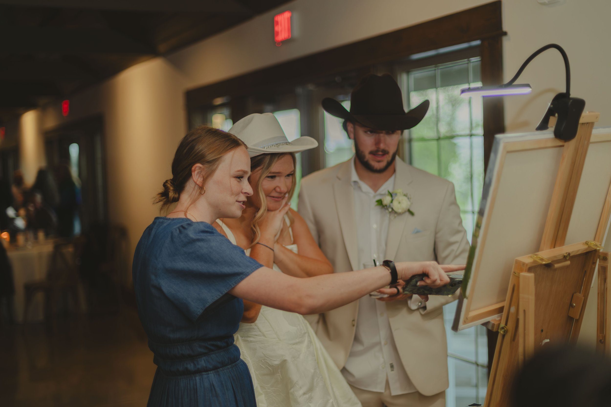 Three people, two women and one man, standing together in front of an easel with a white canvas, looking at something on the canvas. The man is wearing a cowboy hat and a white suit, the woman on the left has red hair and is wearing a blue dress, the woman in the middle has blonde hair and is dressed in white. The scene appears to be at an event or celebration, with a background of indoors setting with windows and a wooden interior.
