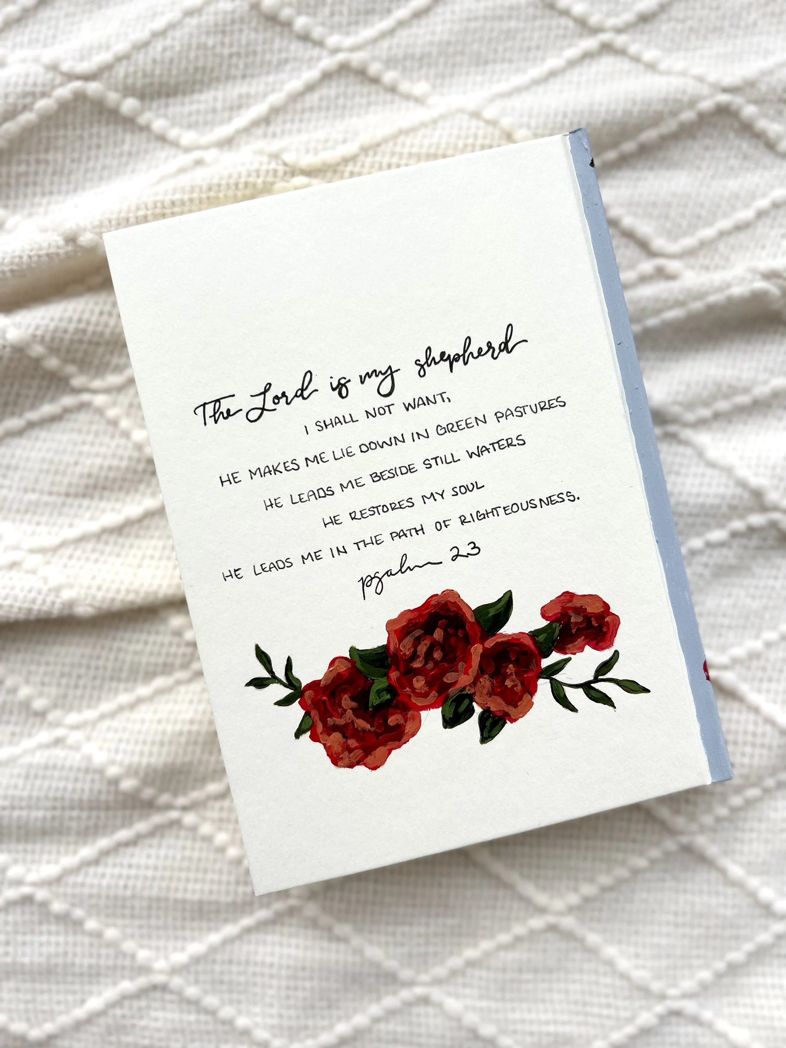 Close-up of a prayer card with a floral illustration of red roses and green leaves, placed on a textured, cream-colored fabric background.