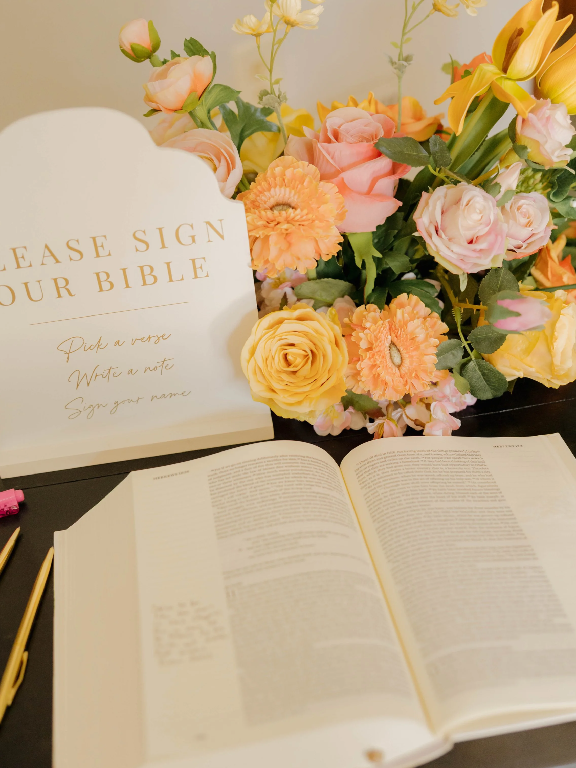 Open book with text, colorful flowers arranged behind it, and a sign that reads 'Please sign your Bible' with space for a verse, note, and name.