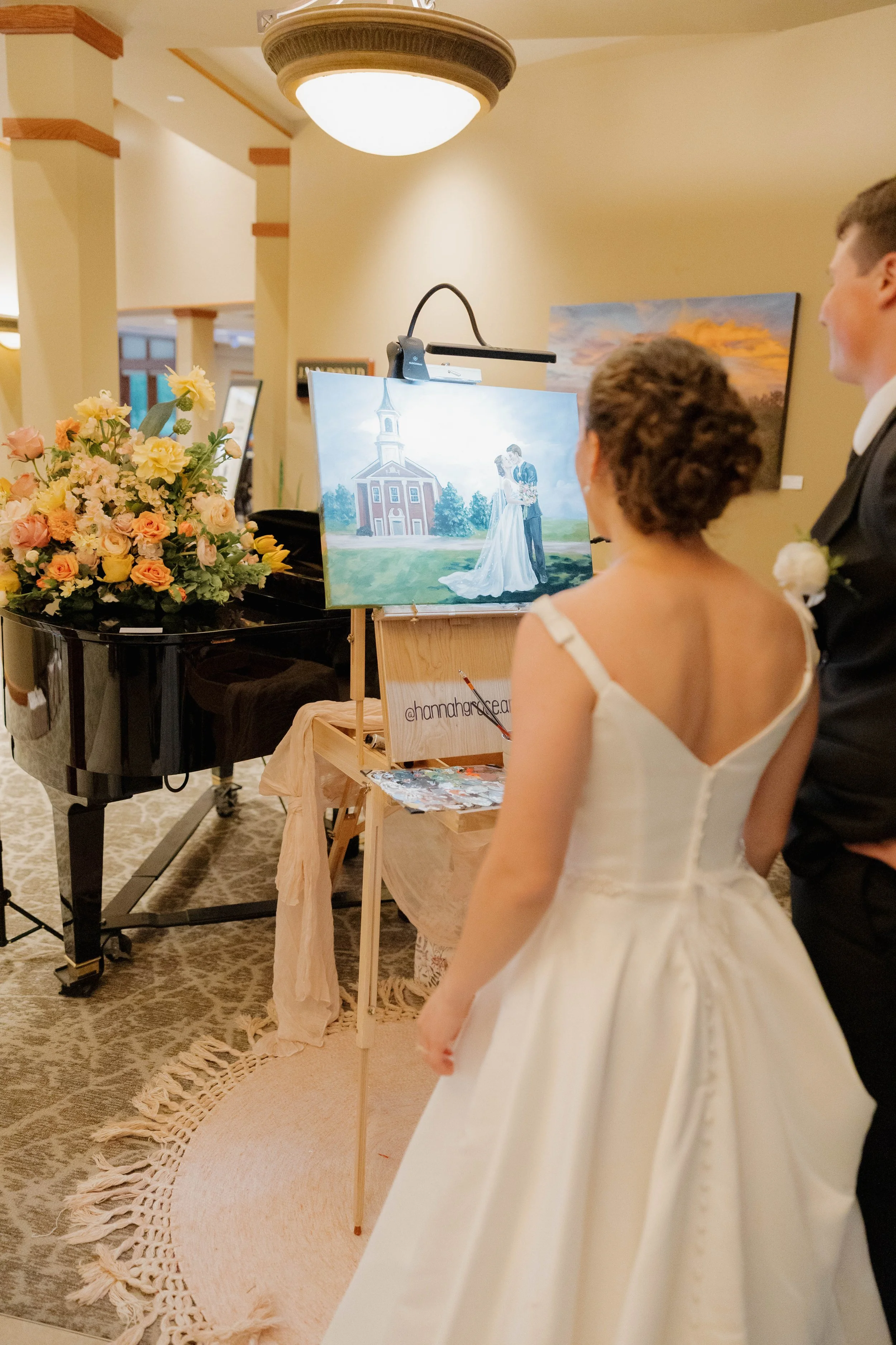 A bride and groom looking at a painting of a wedding scene featuring a church and a couple kissing outside, surrounded by flowers in a wedding or art gallery setting.