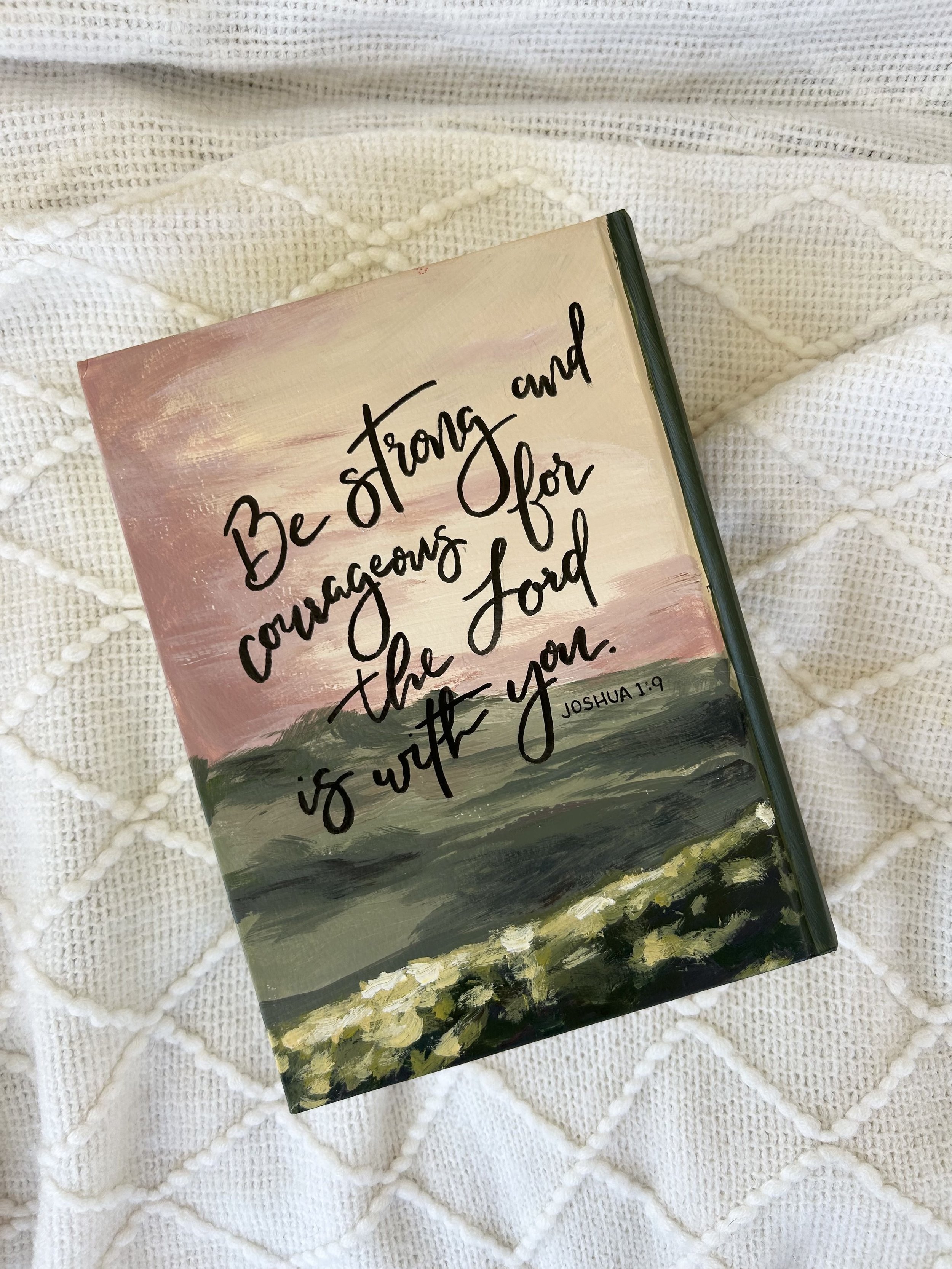 A painted canvas with an inspiring quote from Joshua 1:9 reads 'Be strong and courageous for the Lord is with you,' placed on a white textured knit blanket.