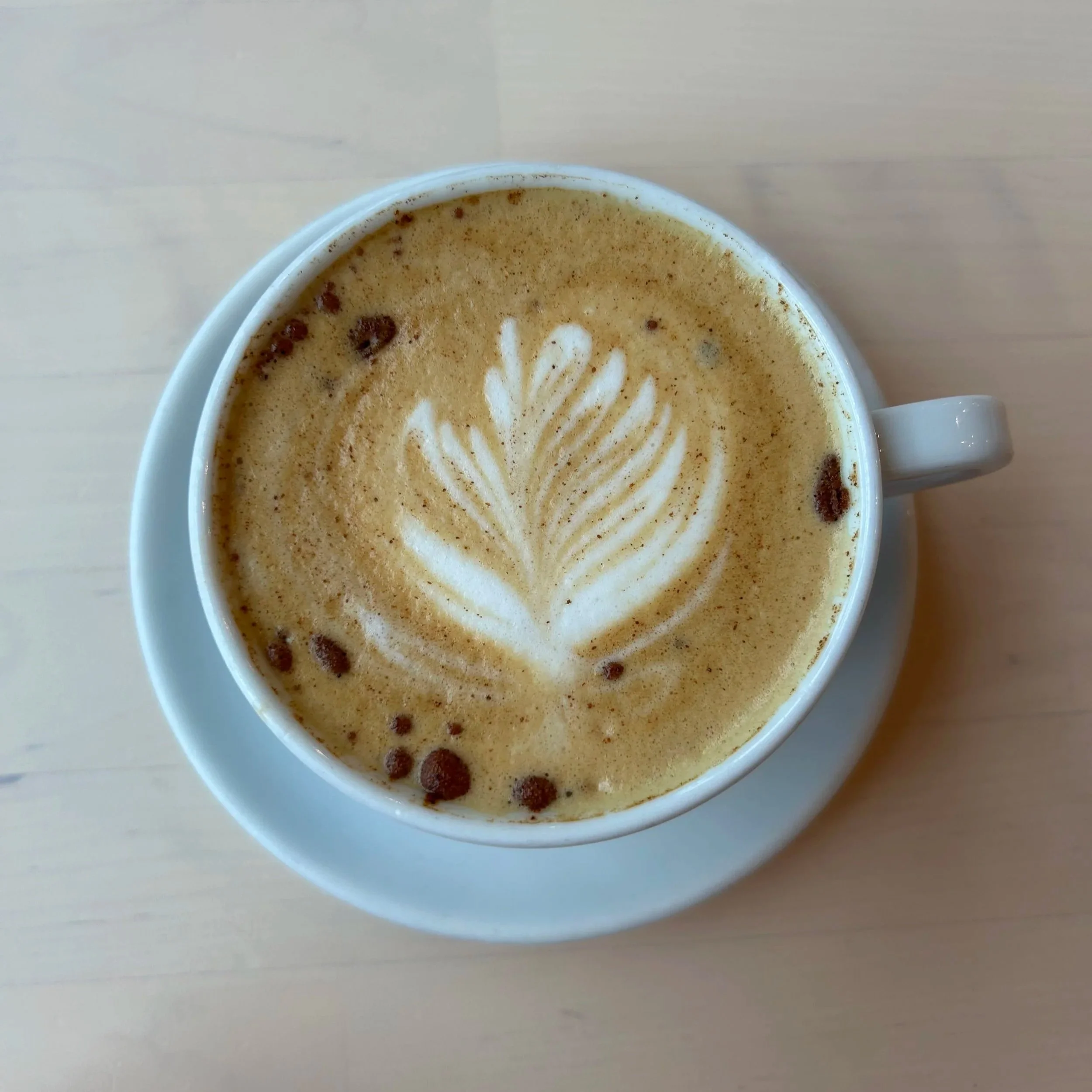 A top view of a cup of coffee with latte art in a white cup and saucer on a light wooden surface.