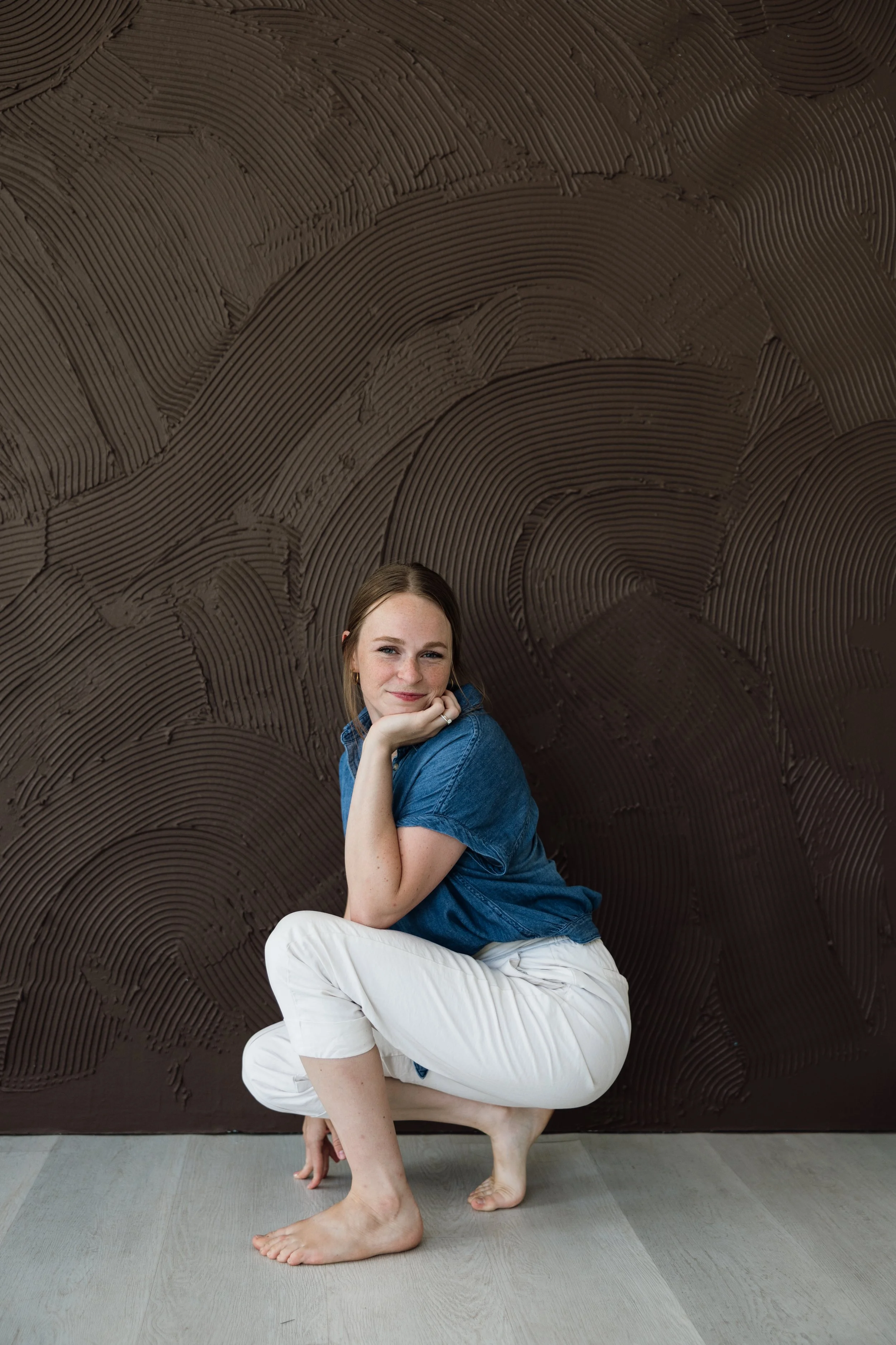 A woman with light skin and brown hair sitting on the floor in front of a textured brown wall. She is wearing a blue shirt and white pants, and is perched in a yoga pose with one knee on the ground and her chin resting on her hand, smiling at the camera.
