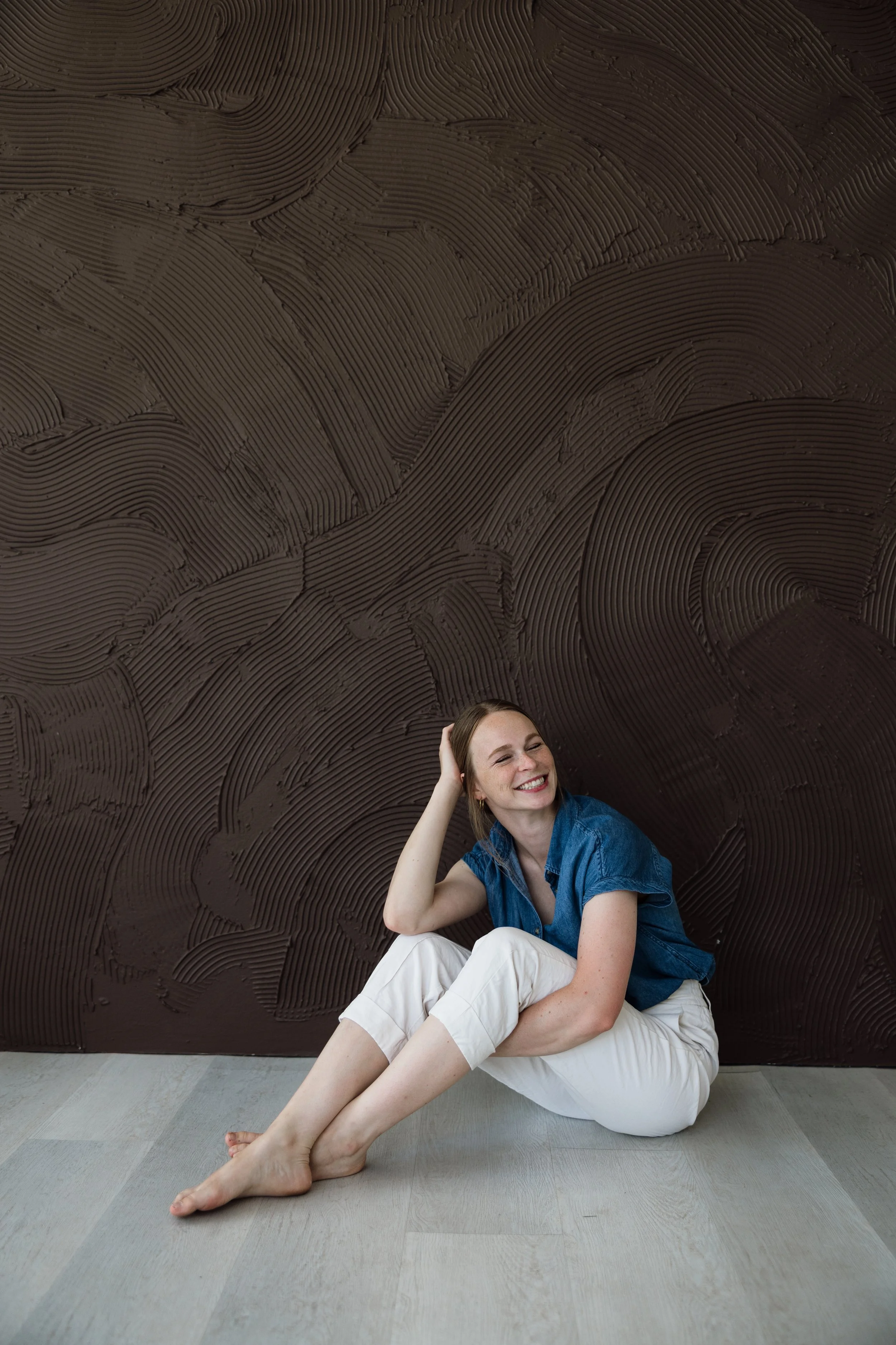 A woman with brown hair smiling and sitting on the floor against a textured dark brown wall. She is wearing a denim shirt and white pants, with her legs extended and her left arm resting on her knee. Her right hand is touching her hair, and she appears relaxed and happy.