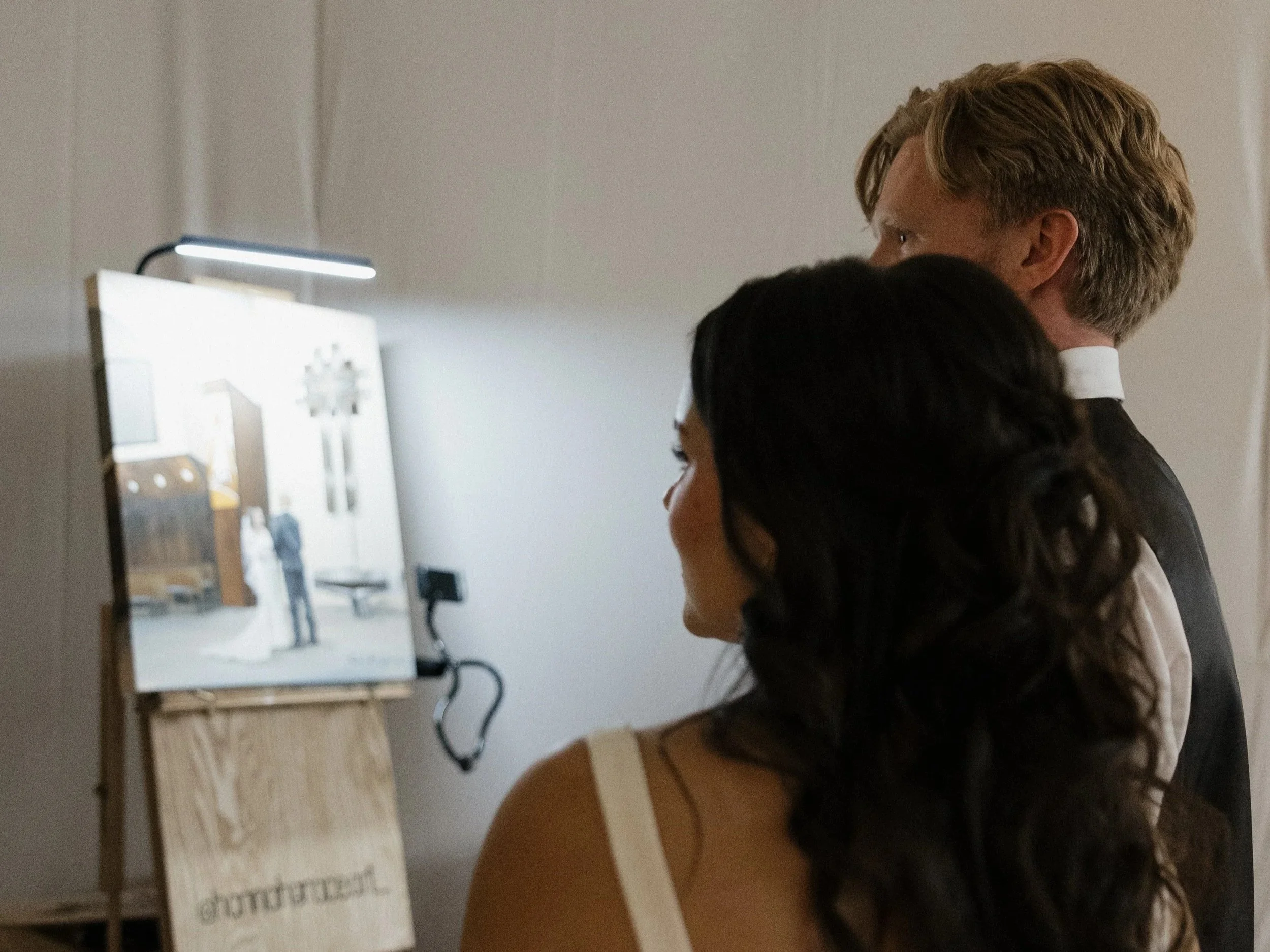 A couple dressed in wedding attire watching a mirror with a bride and groom reflection, with a sign that reads 'Groom & Bride' in front of them.