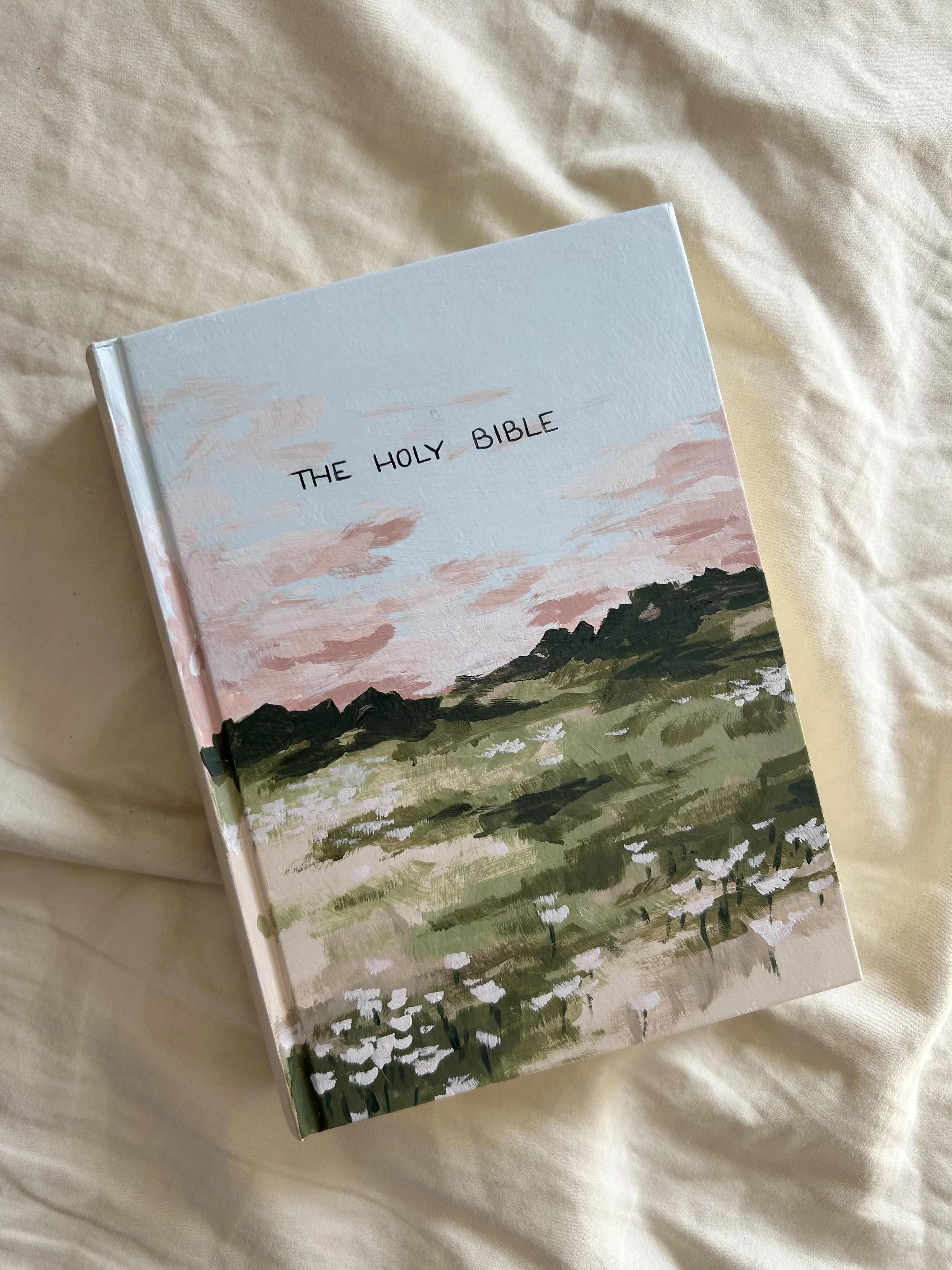 A painting of a landscape with trees and a sky featuring pastel pink and blue hues, titled 'The Holy Bible'.