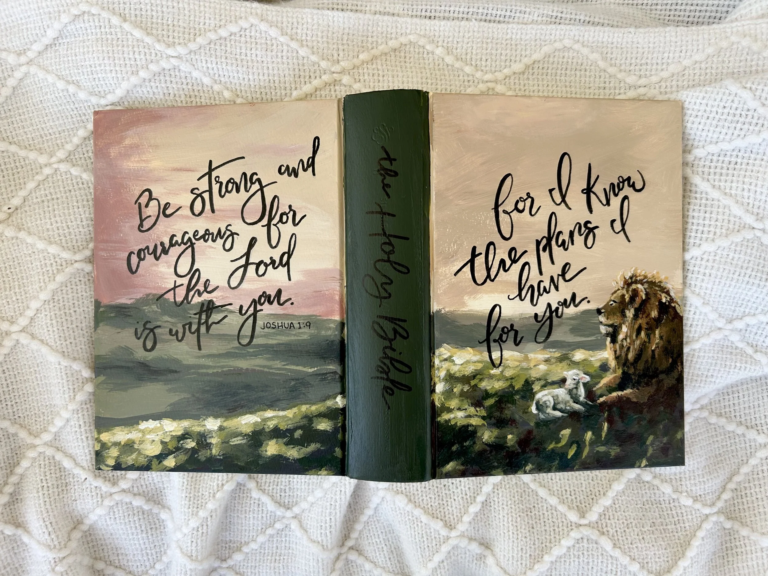 Open hardcover book with painted landscape and handwritten Bible verses on pages, lying on a textured white blanket.