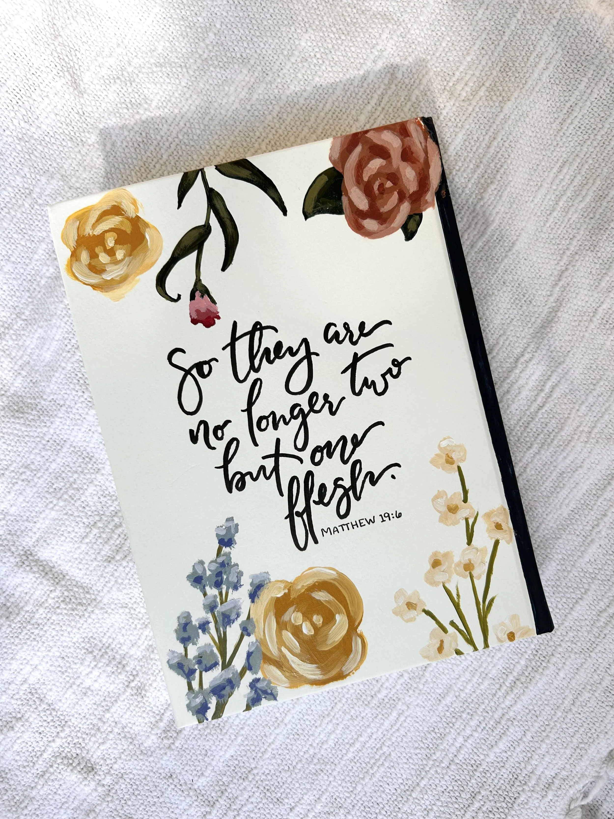 A floral card with painted flowers and a handwritten Bible verse from Matthew 19:6, placed on a white textured fabric.