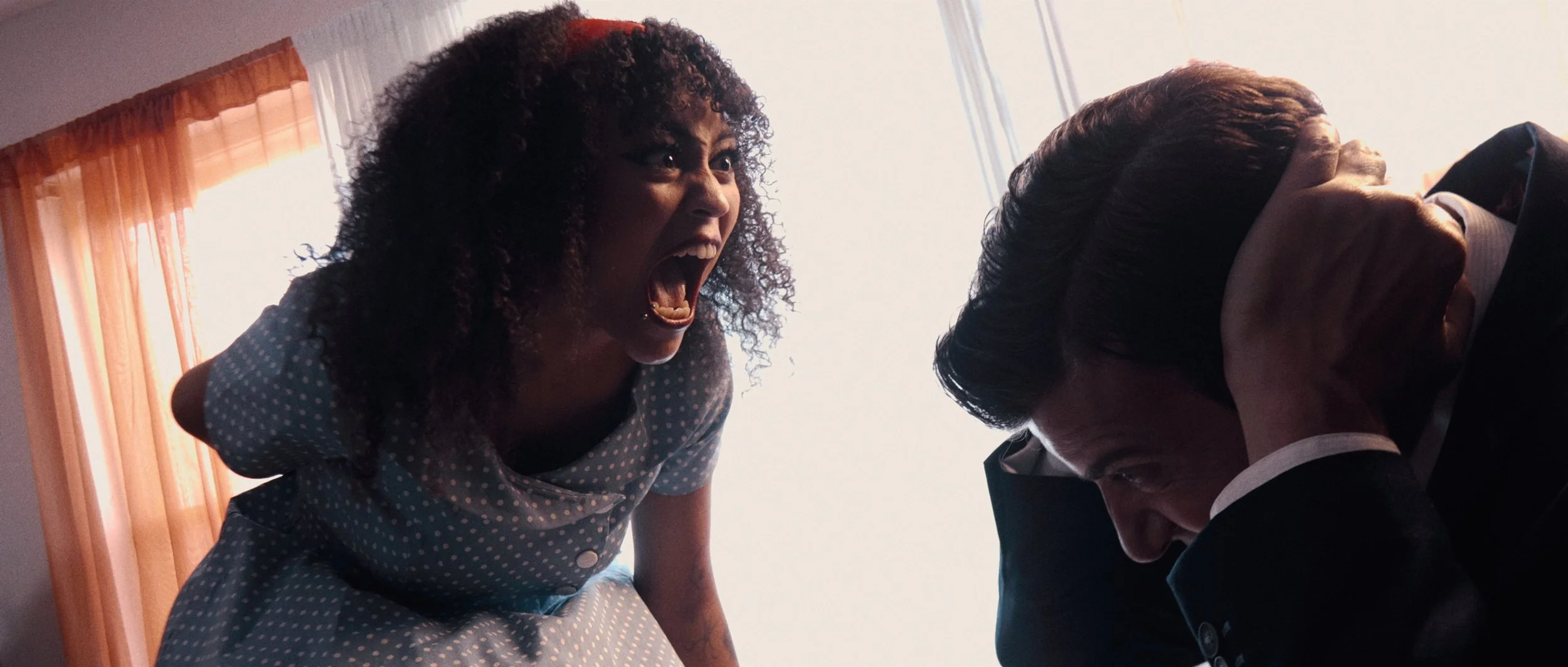 Still from FEMICIDE: a woman in a blue polka-dot dress screams toward a suited man crouching with his hands over his ears.