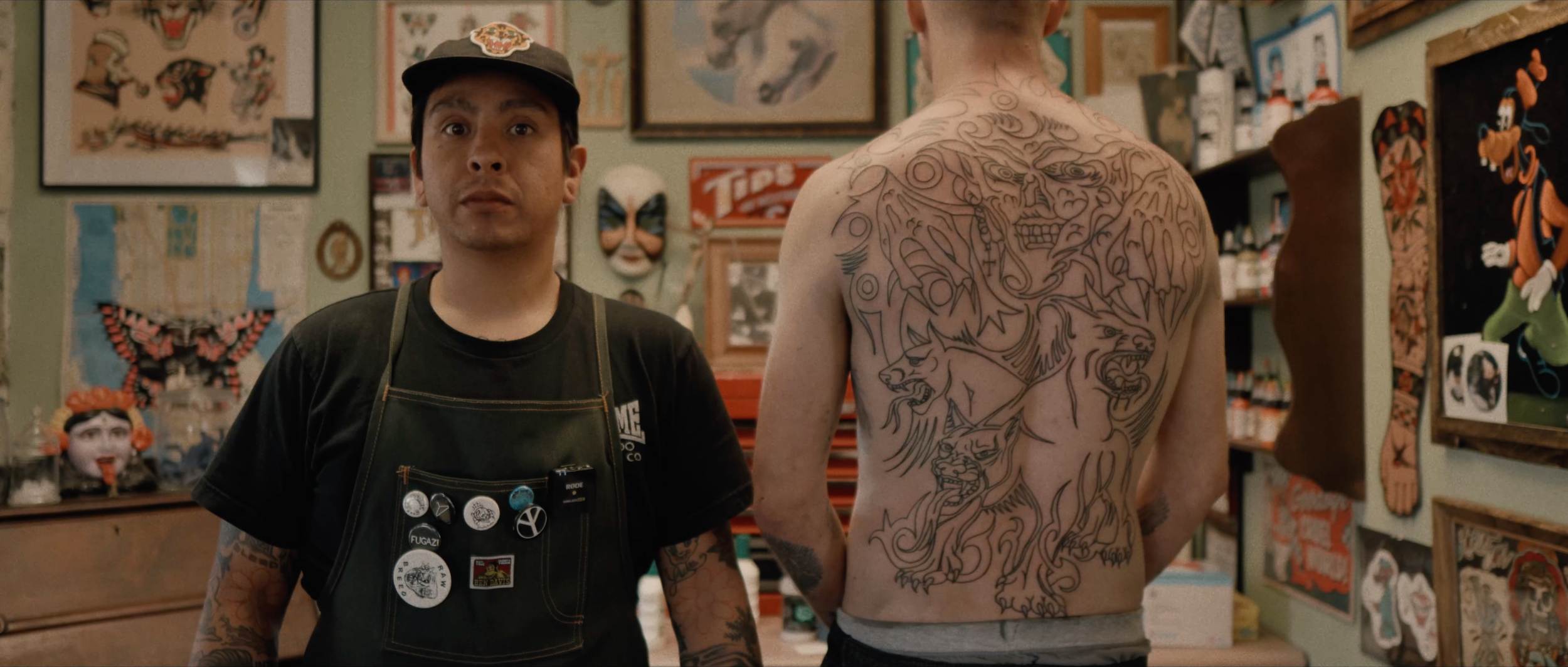 Still from TATTOOER: tattoo artist Joshua Justin Chachi Soto stands beside a client displaying a large back tattoo in an artwork-filled studio.