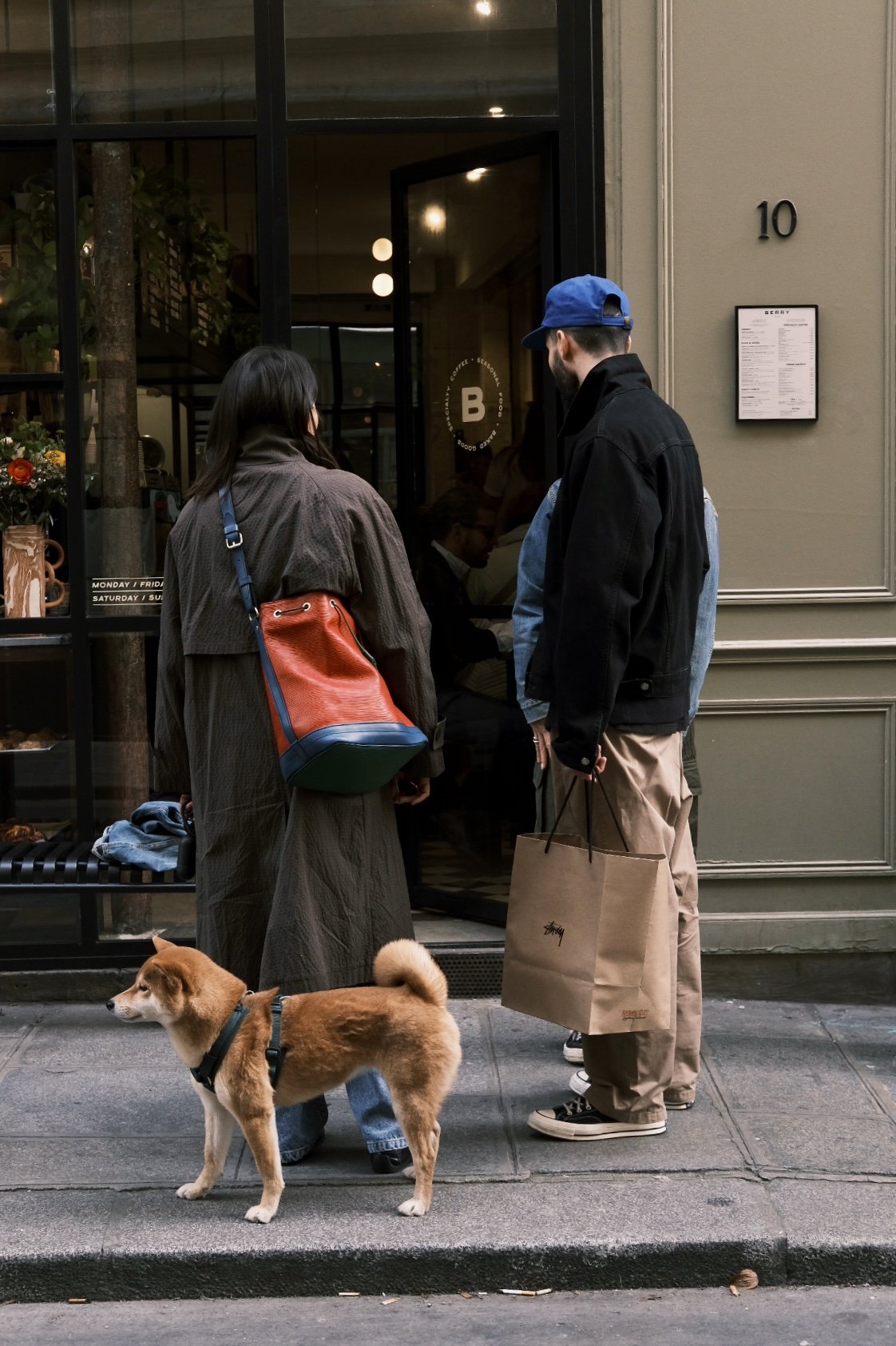 Deux personnes discutent devant une entrée de café ou restaurant, une femme avec un sac rouge, un homme avec un sac en papier, un chien brun avec col bleu à côté, tout en étant à l'extérieur sur le trottoir.