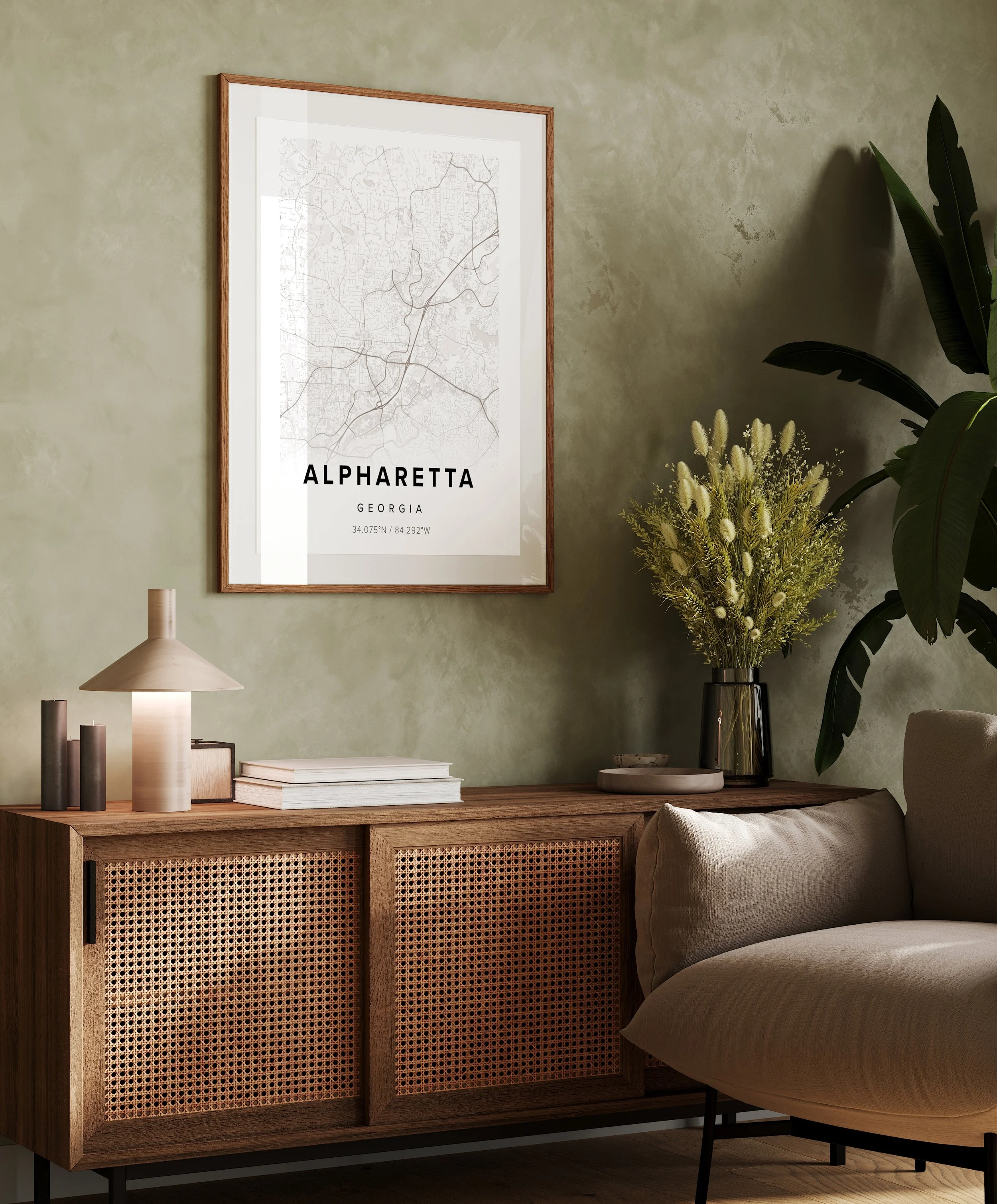 Decorative living room corner with a framed map of Alpharetta, Georgia on a green textured wall, a wooden sideboard with stacked books, a modern table lamp, and a vase with yellow flowers next to a large houseplant and a beige armchair.
