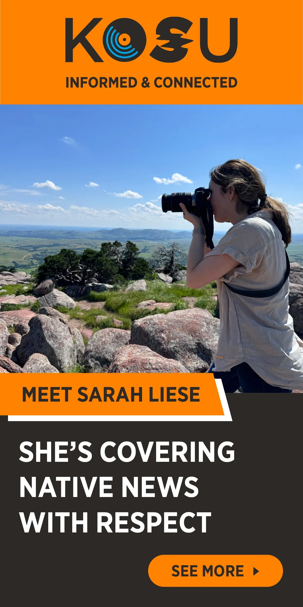 A woman with long hair, wearing a beige shirt, is standing outdoors on rocks, taking a photograph of the landscape with a camera against a blue sky with scattered clouds. The background shows green hills and trees.