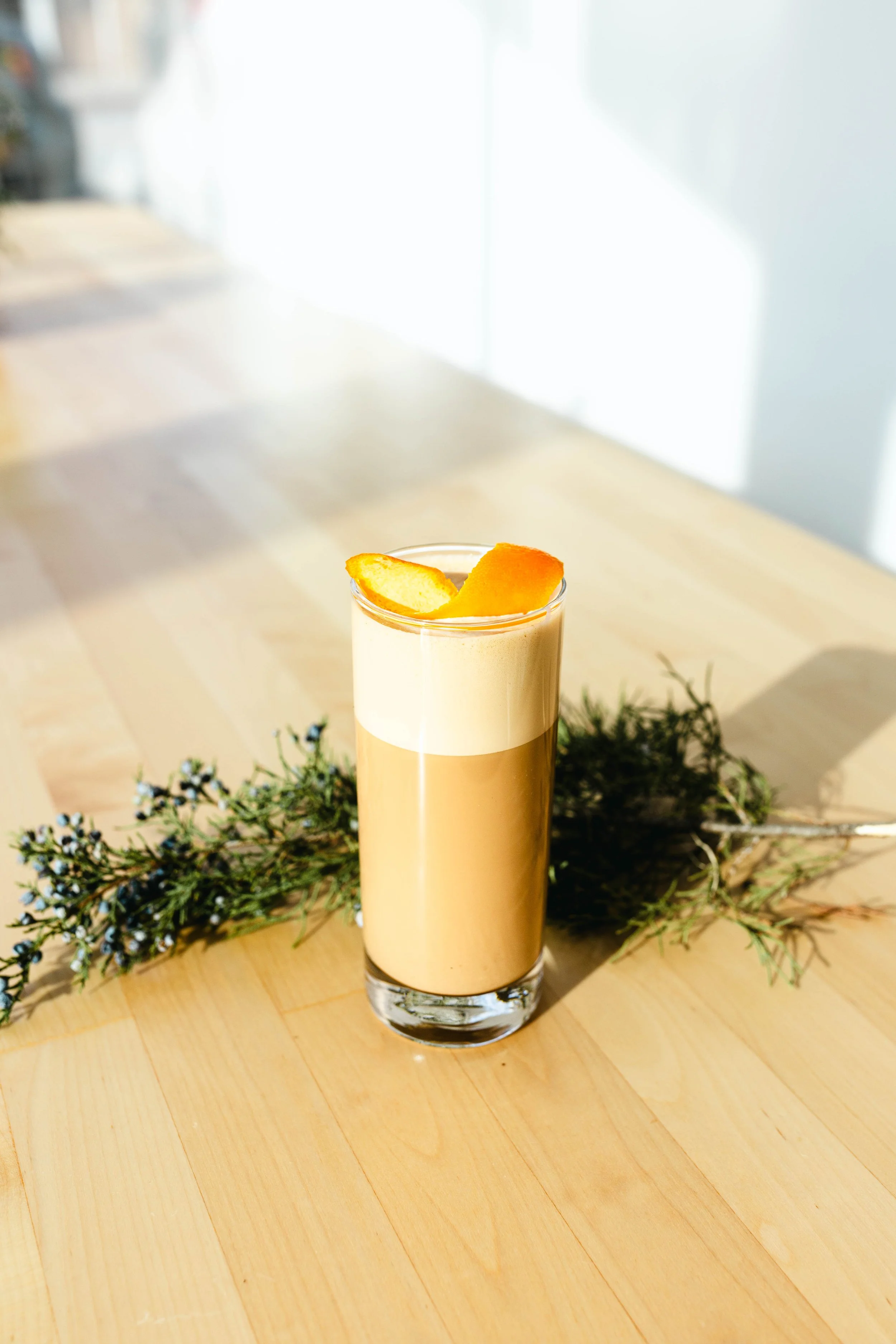 Latte in a tall glass with orange garnish on a wooden table with greenery.