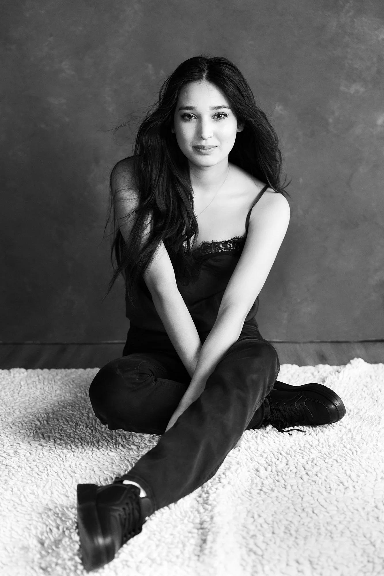 Black and white portrait of a young woman sitting cross-legged on a white rug, gazing softly into the camera with a calm and open expression.