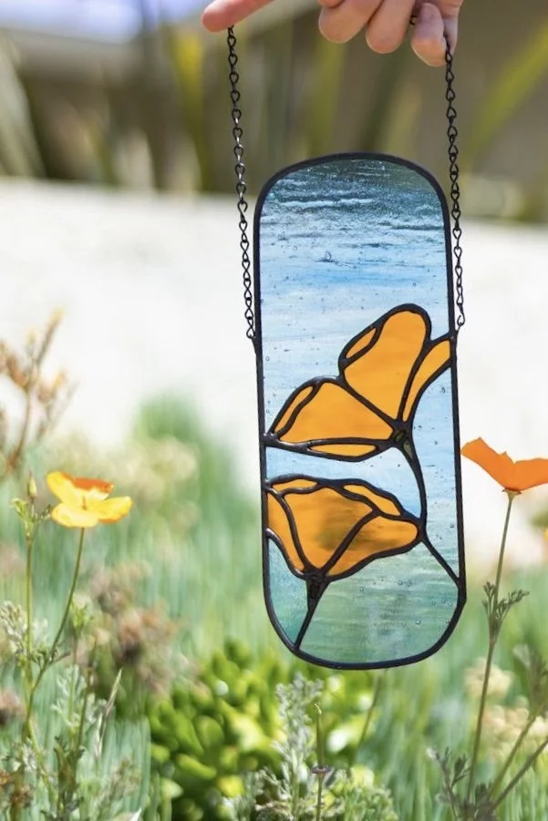 Oval-shaped stained glass panel featuring bright orange California poppies surrounded by sky blue textured glass