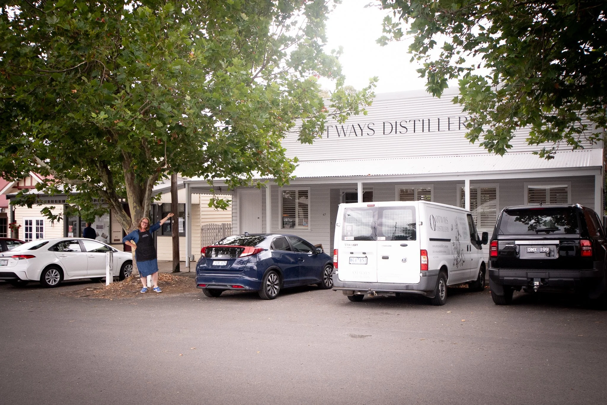 A woman wearing a black apron pointing at a large sign for 'Five Ways Distillery' with parked cars and trees in the background.