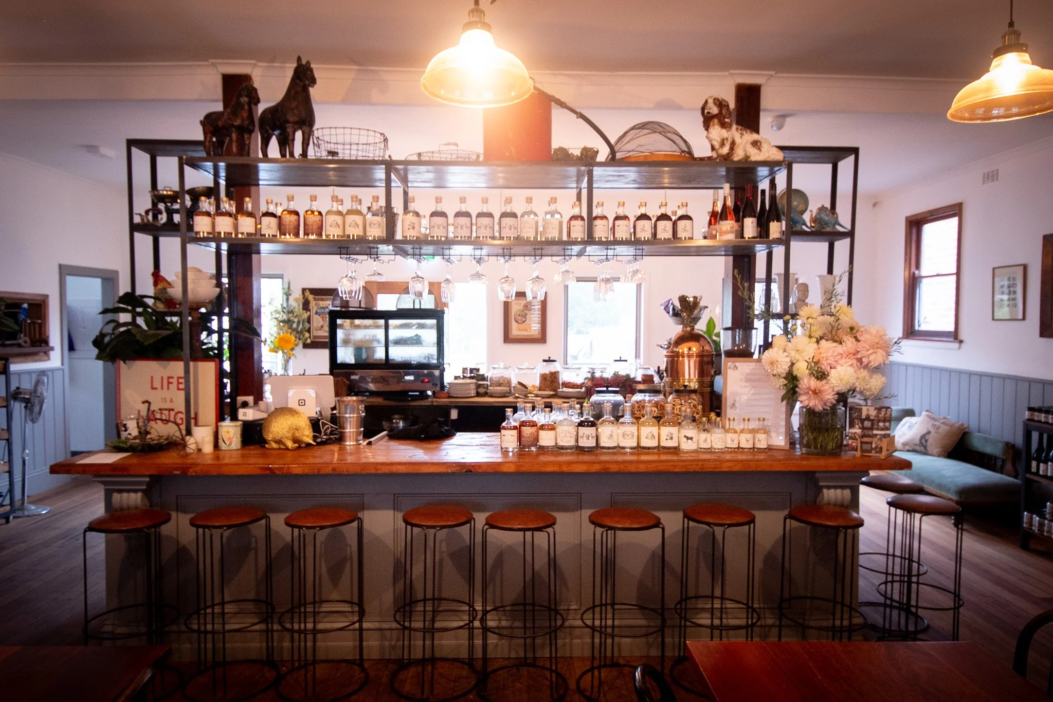 A bar with shelves of liquor bottles, bar stools, and decorative items including flowers, a zebra figurine, and horse statues on top of the shelves. Soft lighting and windows in the background create a cozy atmosphere.