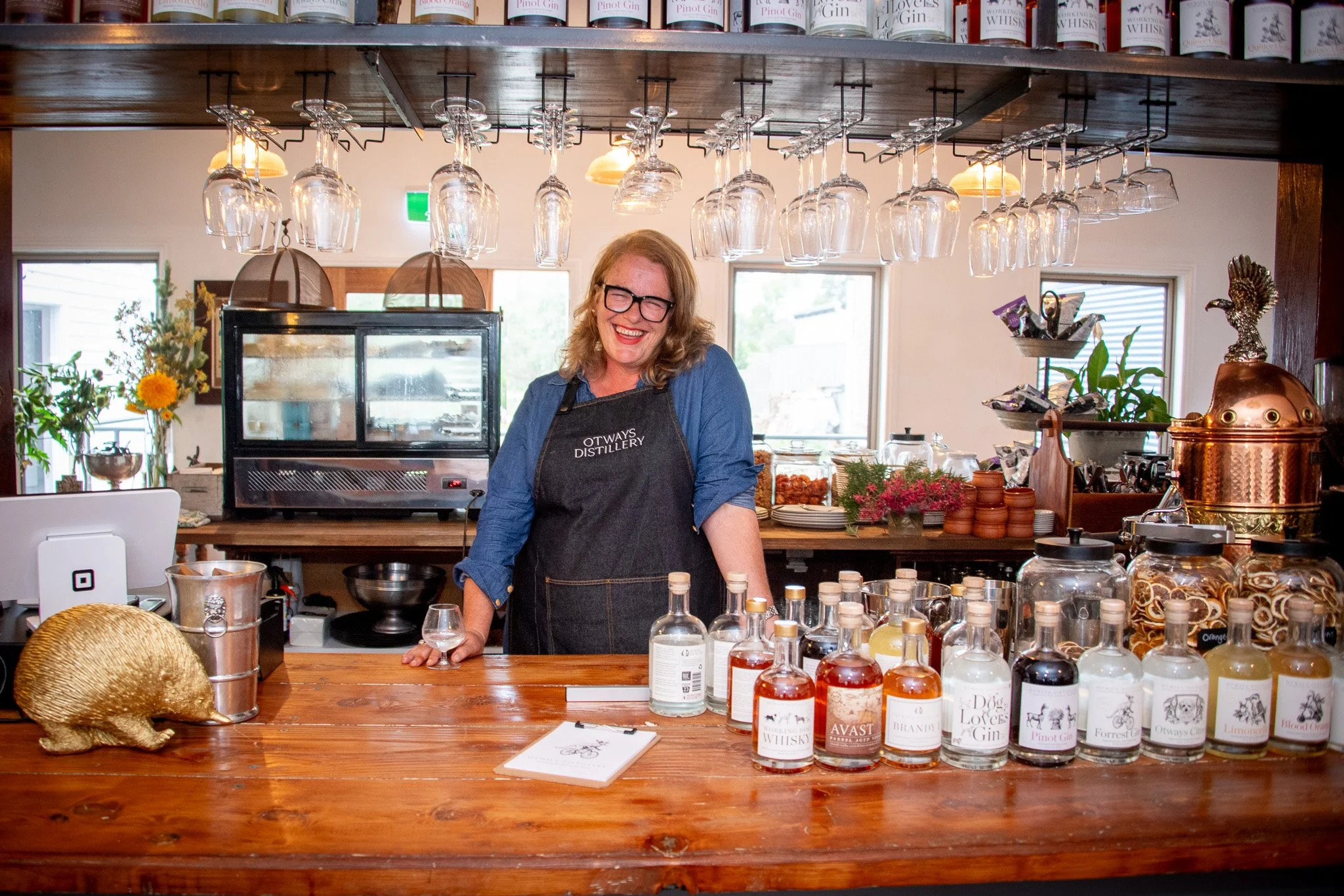 A woman with glasses and curly hair wearing a black apron smiling behind a bar counter with various bottles of flavored spirits, glasses hanging above, and decorative items like a gold rabbit sculpture and flowers, in a cozy bar or tasting room setti