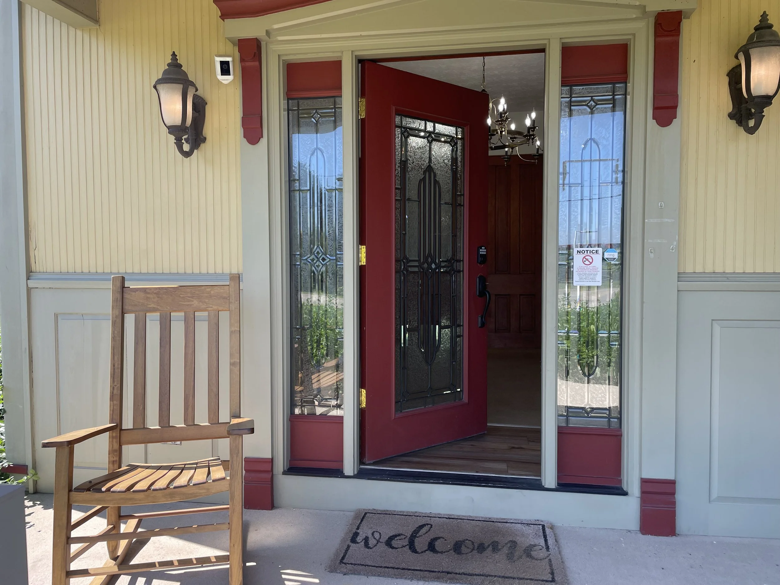 Front porch of a house with a red door slightly open, a wooden chair to the left, a welcome doormat, two wall-mounted lantern lights, and glass panel sidelights.