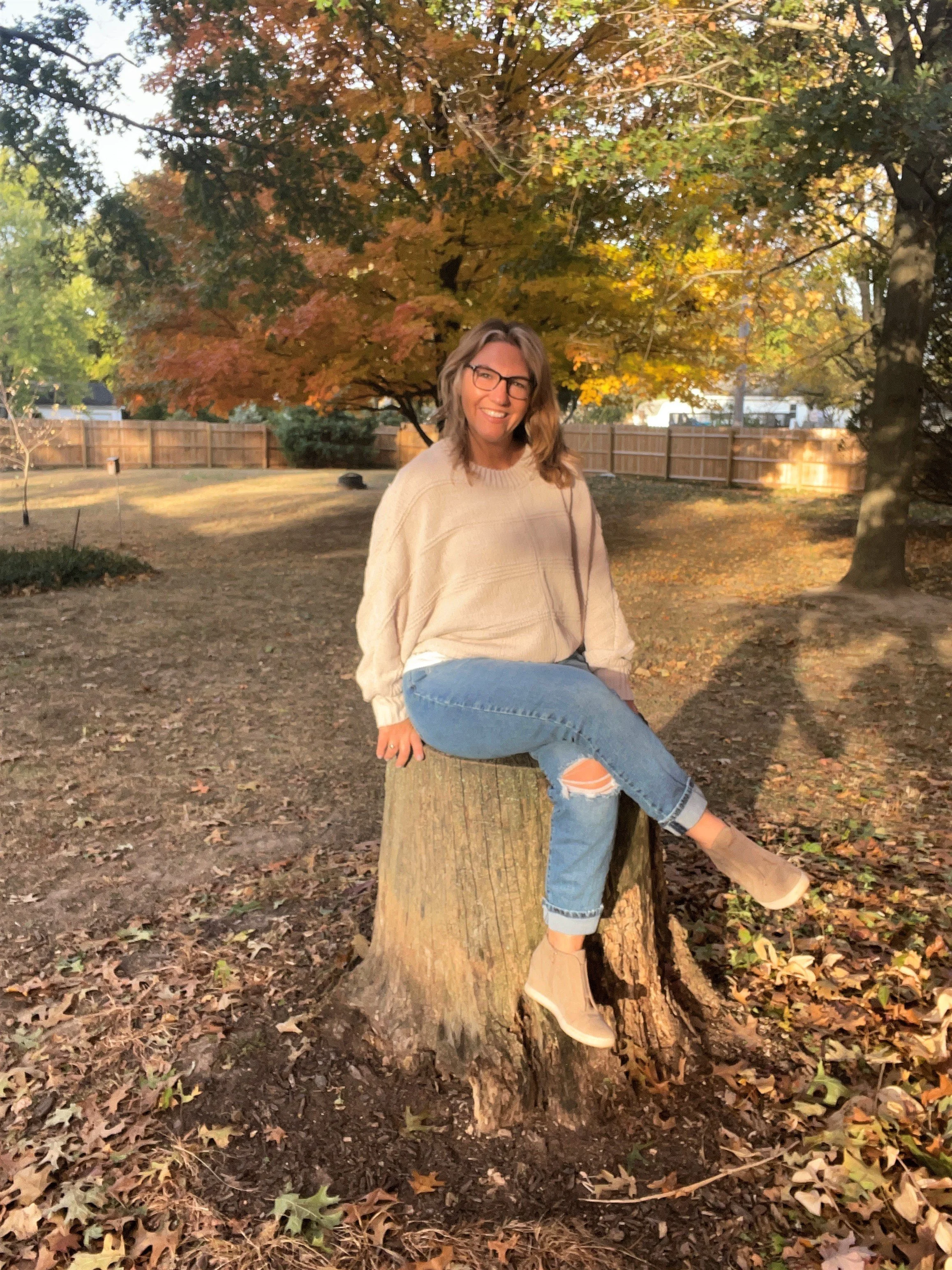 a woman sitting on a stump in the sun with a tree with fall leaves on it behind her