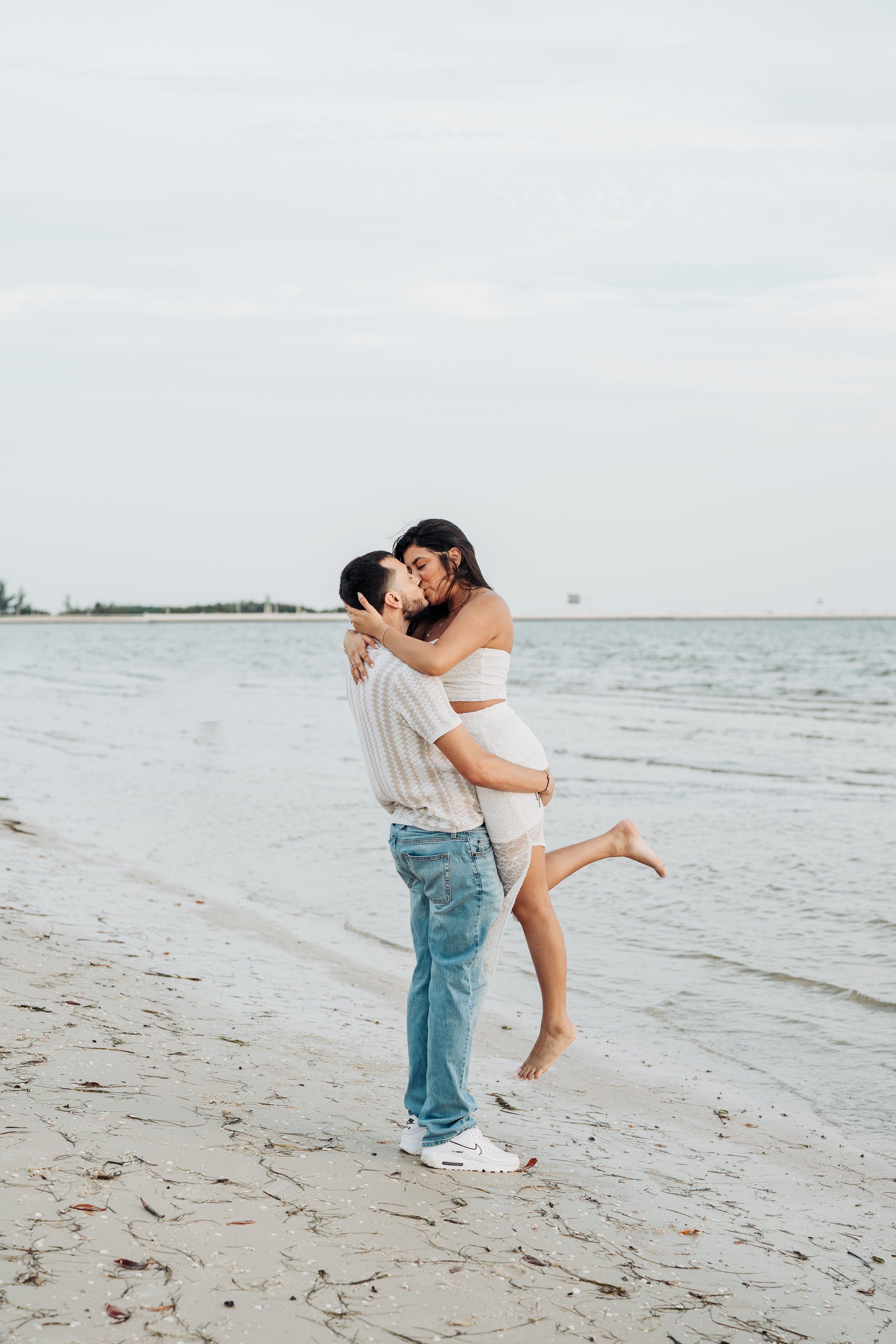 A couple is standing on a beach near the water, with the man lifting the woman in his arms as they kiss. The woman has her arms around the man's head, and they are both smiling, enjoying a romantic moment by the ocean under a cloudy sky.