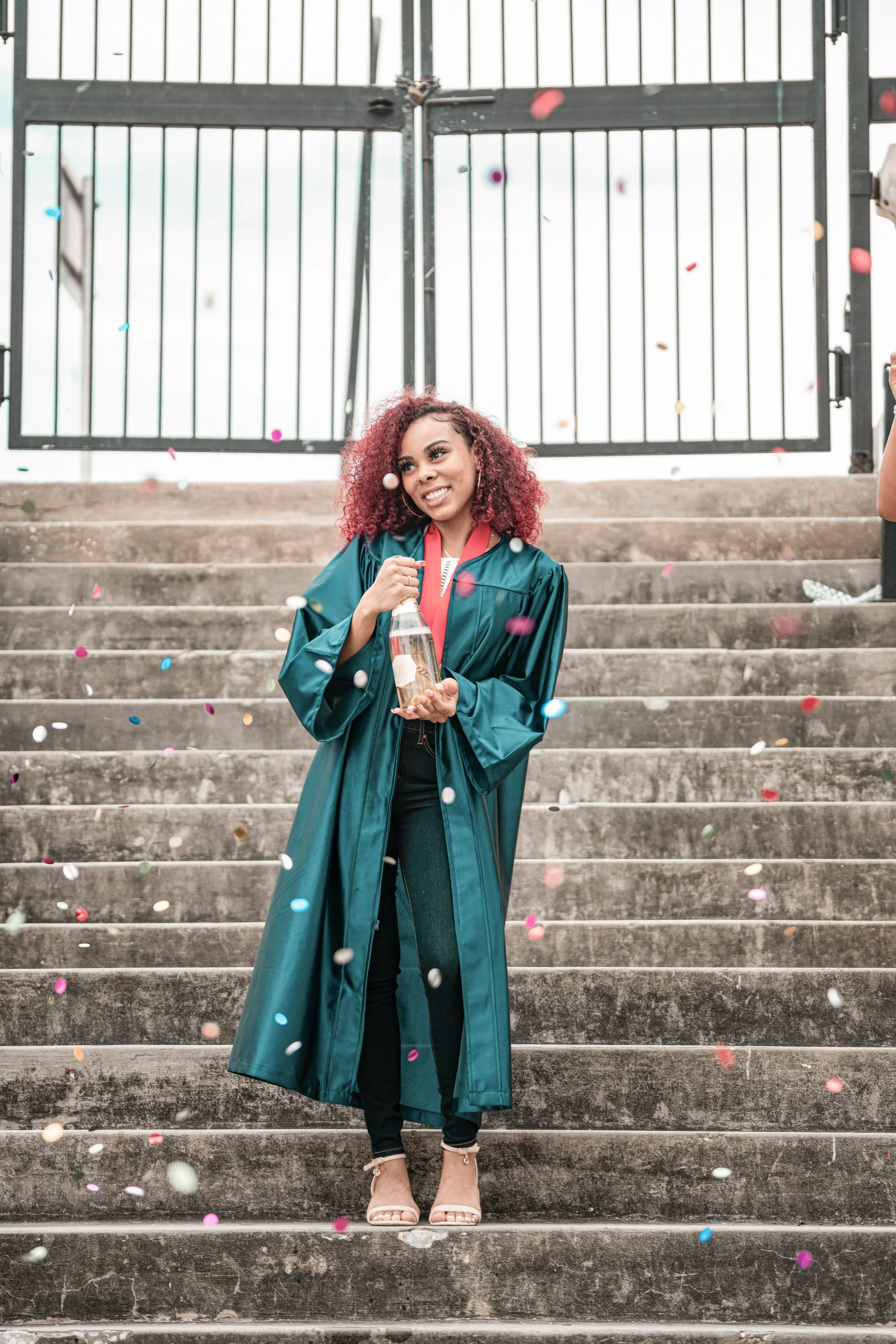 Young woman in a teal graduation gown and heels holding a bottle, standing on concrete stairs in front of a gated fence, with colorful confetti falling around her.