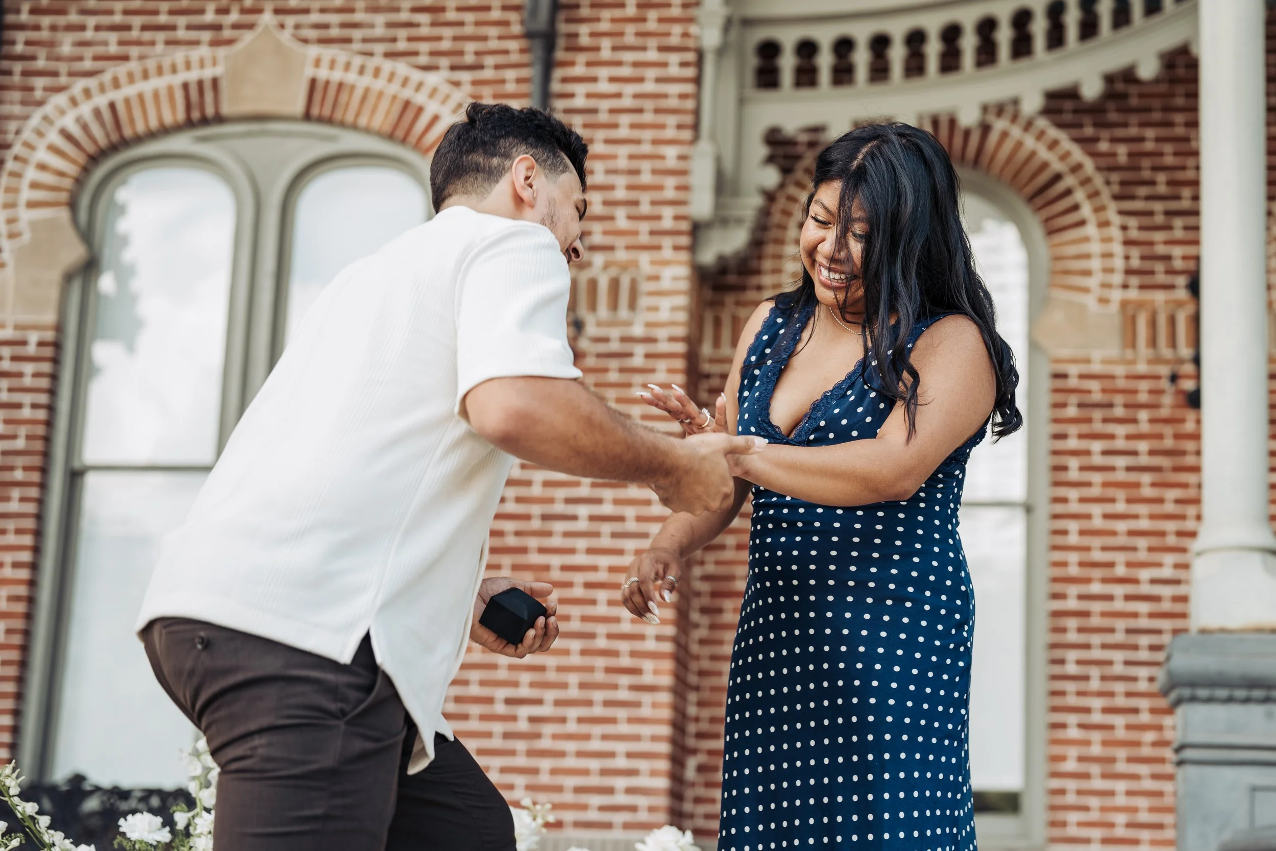 A man in a white shirt appears to be proposing marriage to a woman in a blue polka dot dress in front of a brick building, with both of them smiling and holding hands.