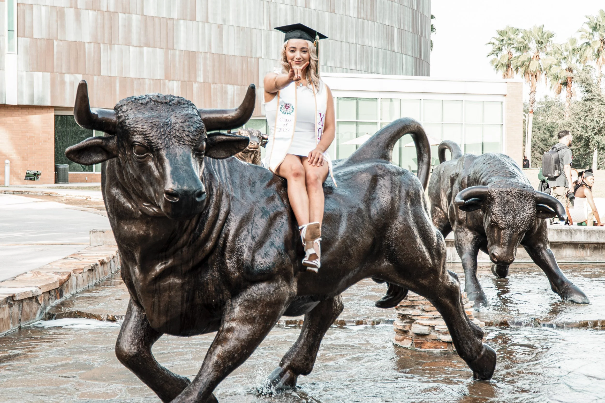 Young woman in graduation cap and gown sitting on a large bronze bull statue in an outdoor plaza, pointing towards the camera, with modern building and palm trees in the background.