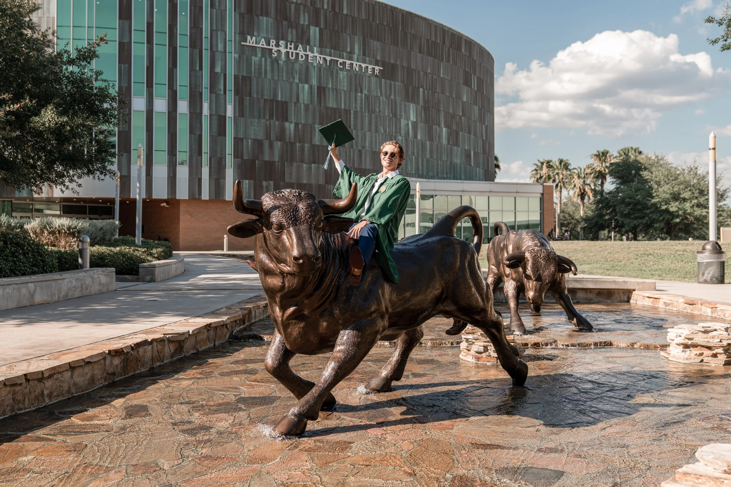 A graduate in a green cap and gown sitting on a sculpture of a buffalo at Marshall Student Center with a modern building and trees in the background.