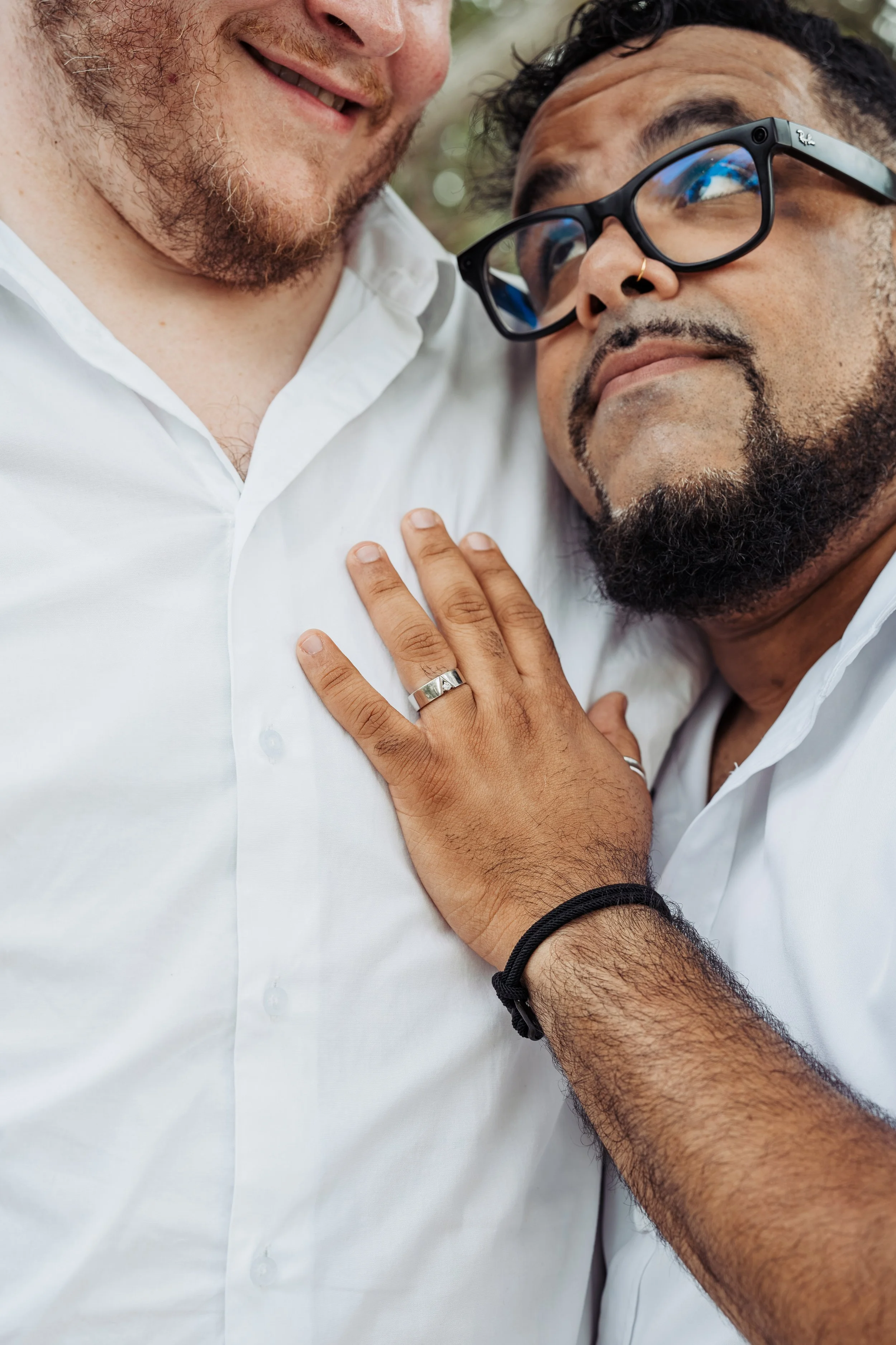 Close-up of two men with one man touching the other's chest. The man on the right wears glasses and a nose ring, and the man on the left has a beard and a white shirt.