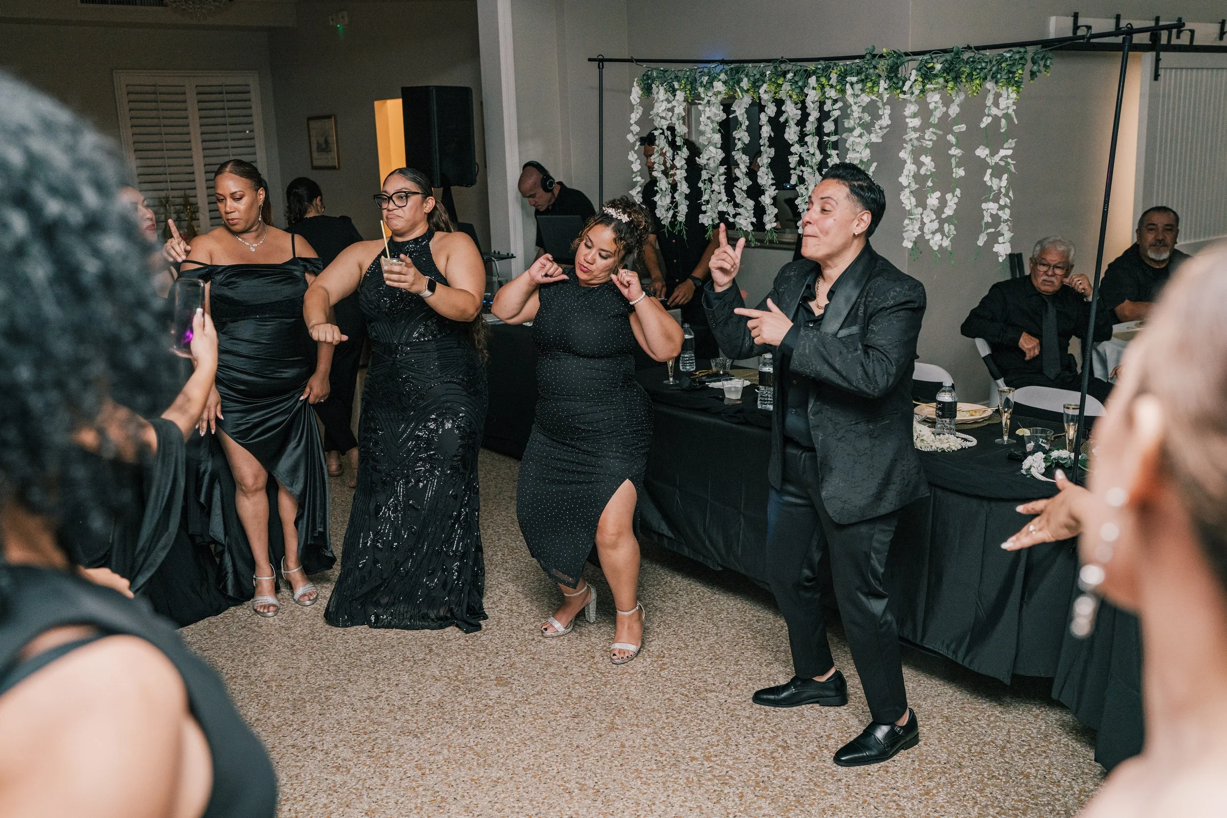 People dancing at a party or celebration, with a floral backdrop in the background.