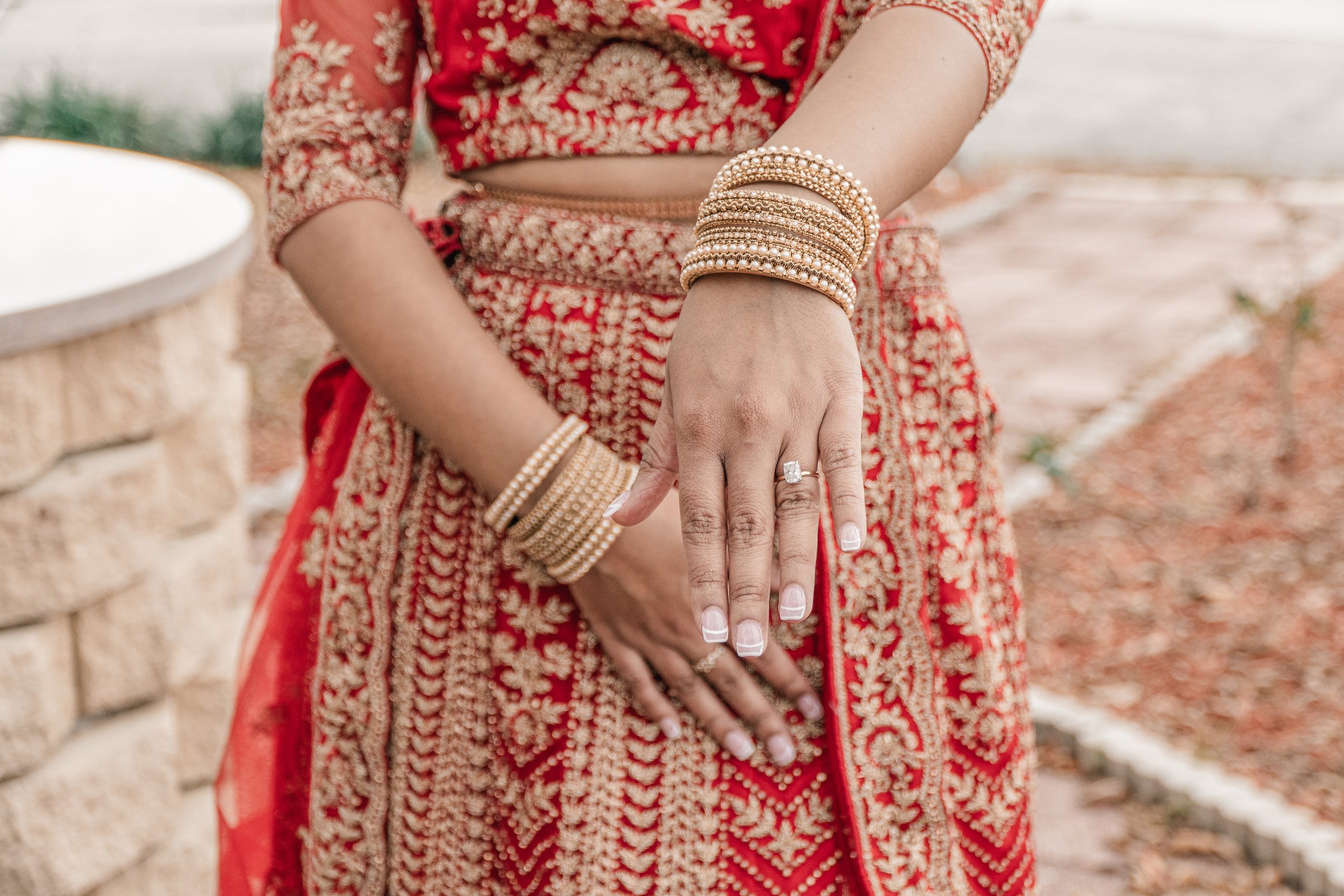 Close-up of a woman wearing traditional red and gold Indian attire with gold and pearl bracelets and a large ring, standing near a stone wall.