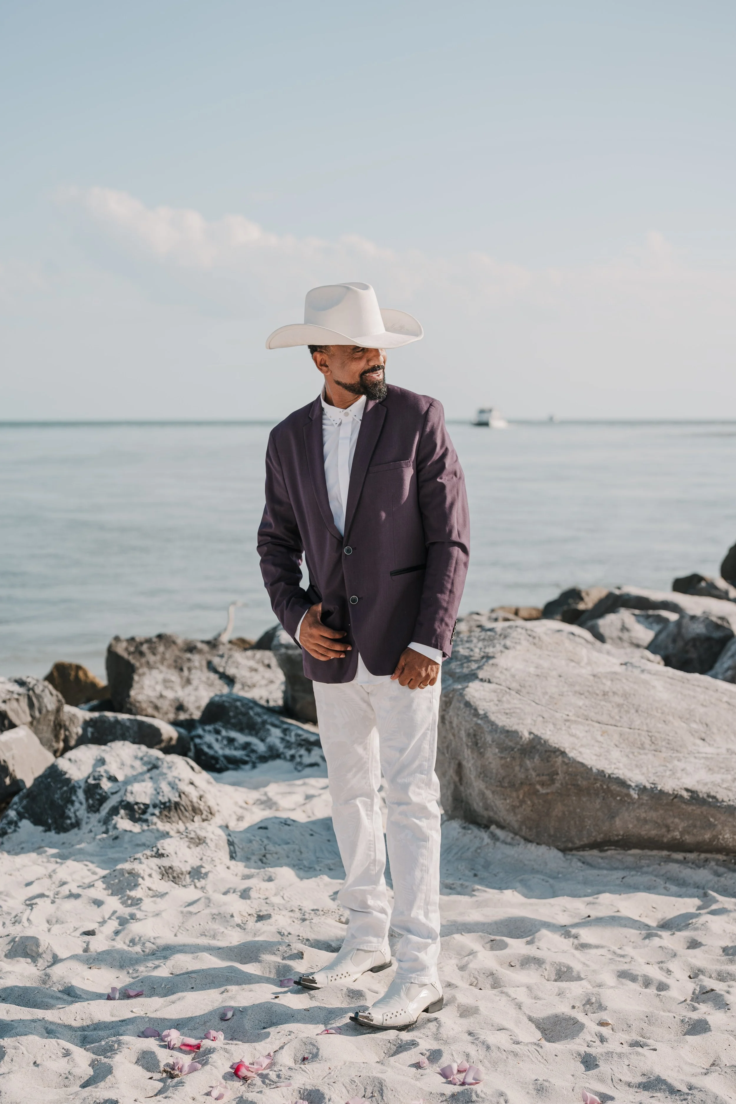 Man wearing a large white cowboy hat, purple blazer, white shirt, white pants, and white shoes standing on a beach with rocks and pink flower petals on the sand, with the ocean and a boat in the background.