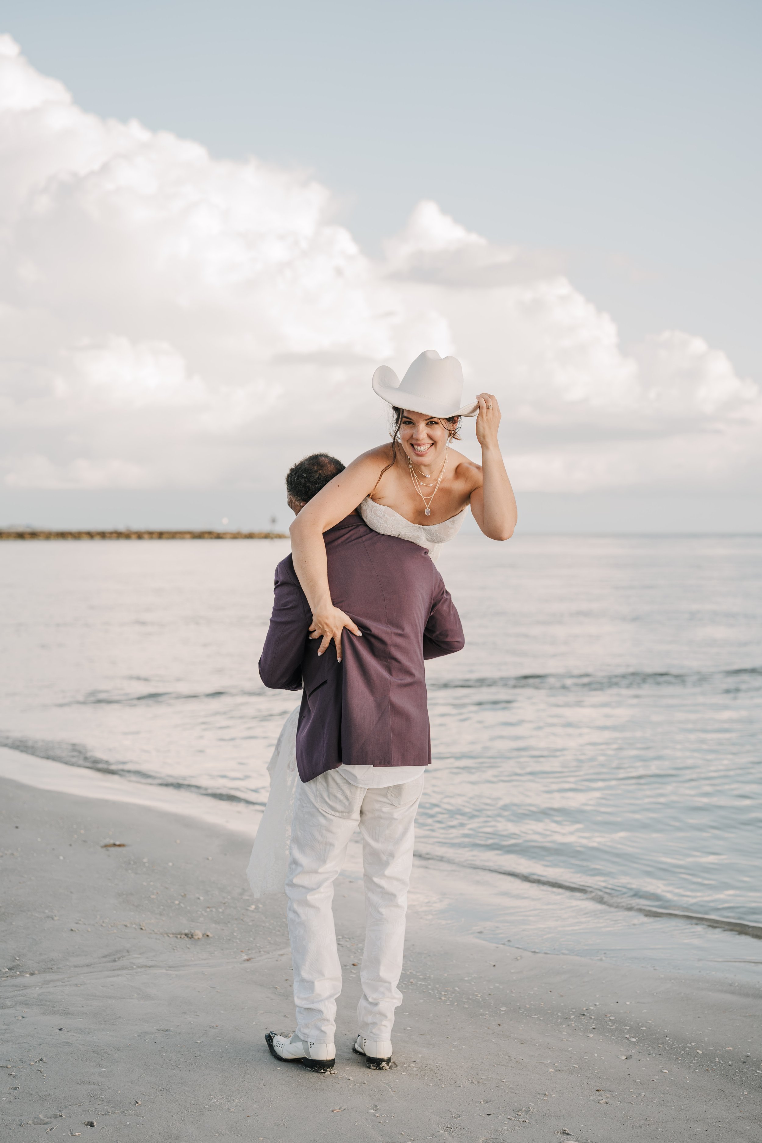A woman with a wide-brimmed white hat and layered jewelry, smiling, on a beach, being lifted by a person in a suit, with the ocean and cloudy sky in the background.