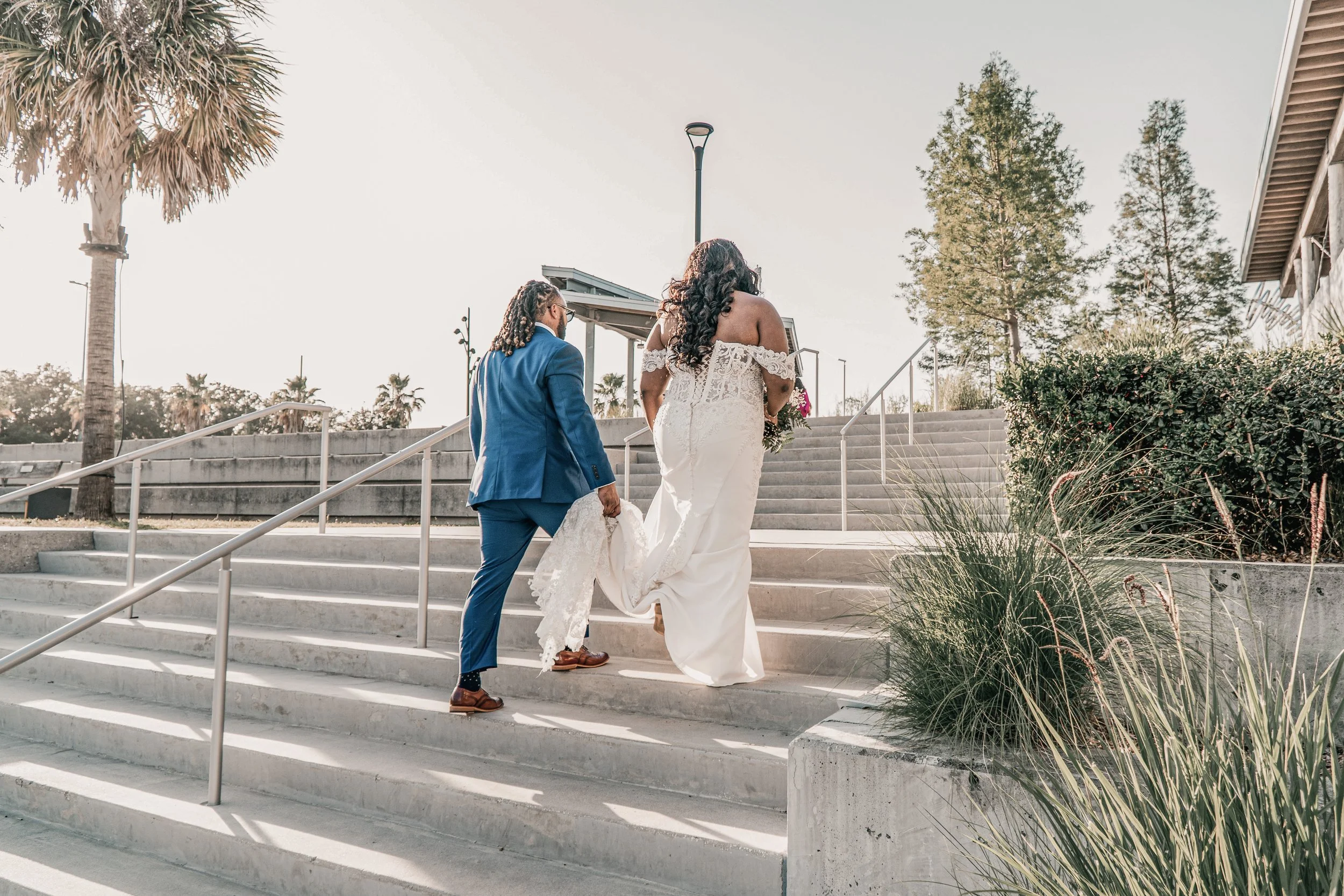 A couple, a man in a blue suit and a woman in a white wedding dress, walking up outdoor stairs in a park-like setting during daylight, with trees and plants surrounding the area.