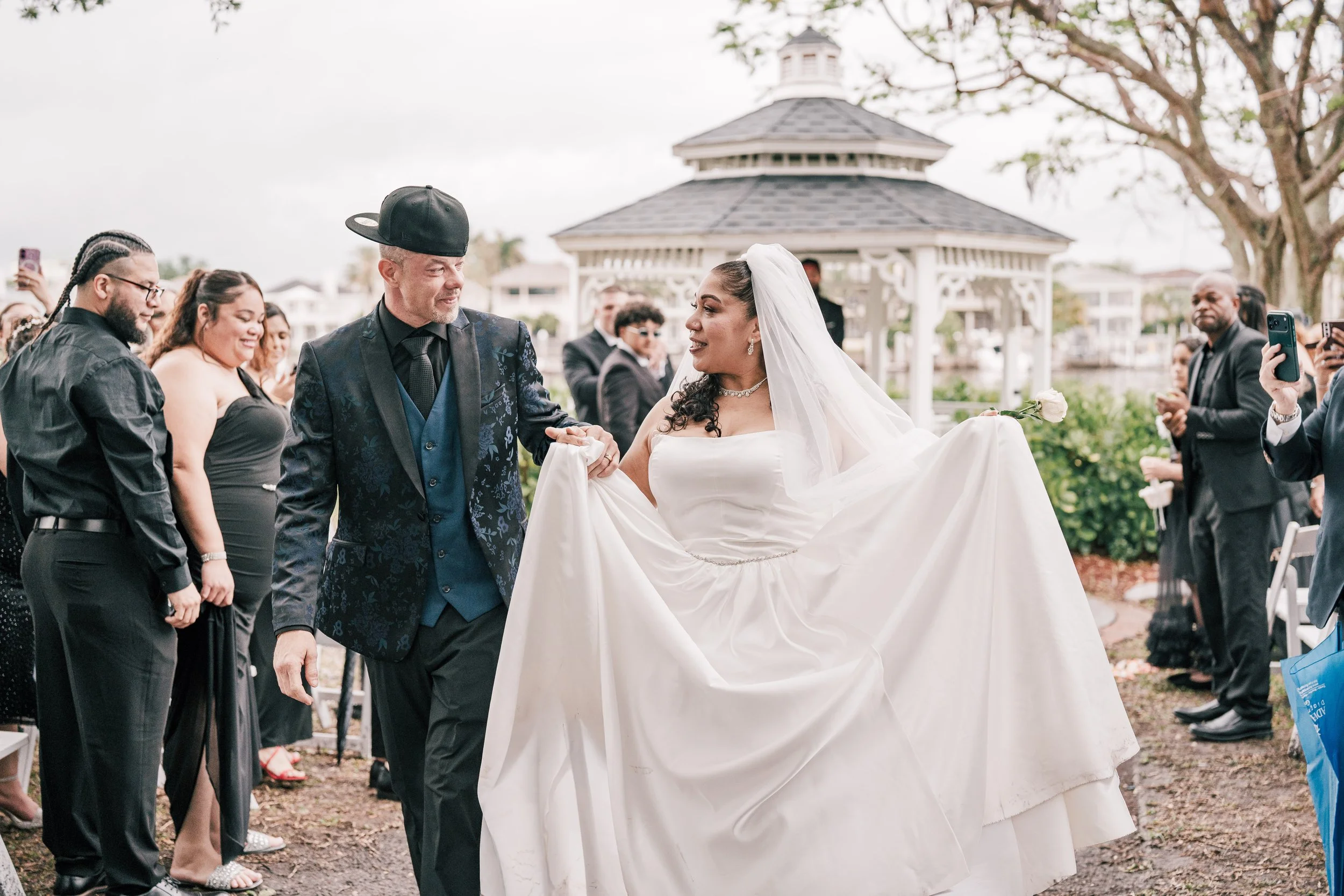 Bride and groom holding hands during their wedding ceremony, surrounded by guests outdoors with a gazebo in the background.