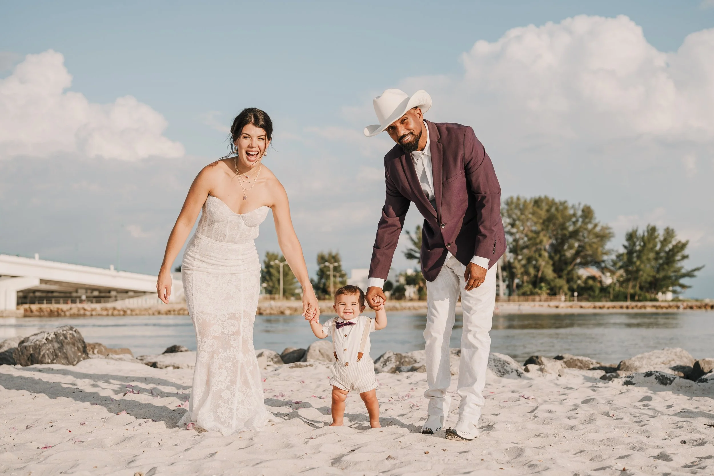 A family of four enjoying a day at the beach, including a woman in a white dress, a man in a burgundy blazer and white hat, a toddler in a beige outfit, and a young girl in a white dress with a bowtie, standing on the sand near the water under partly