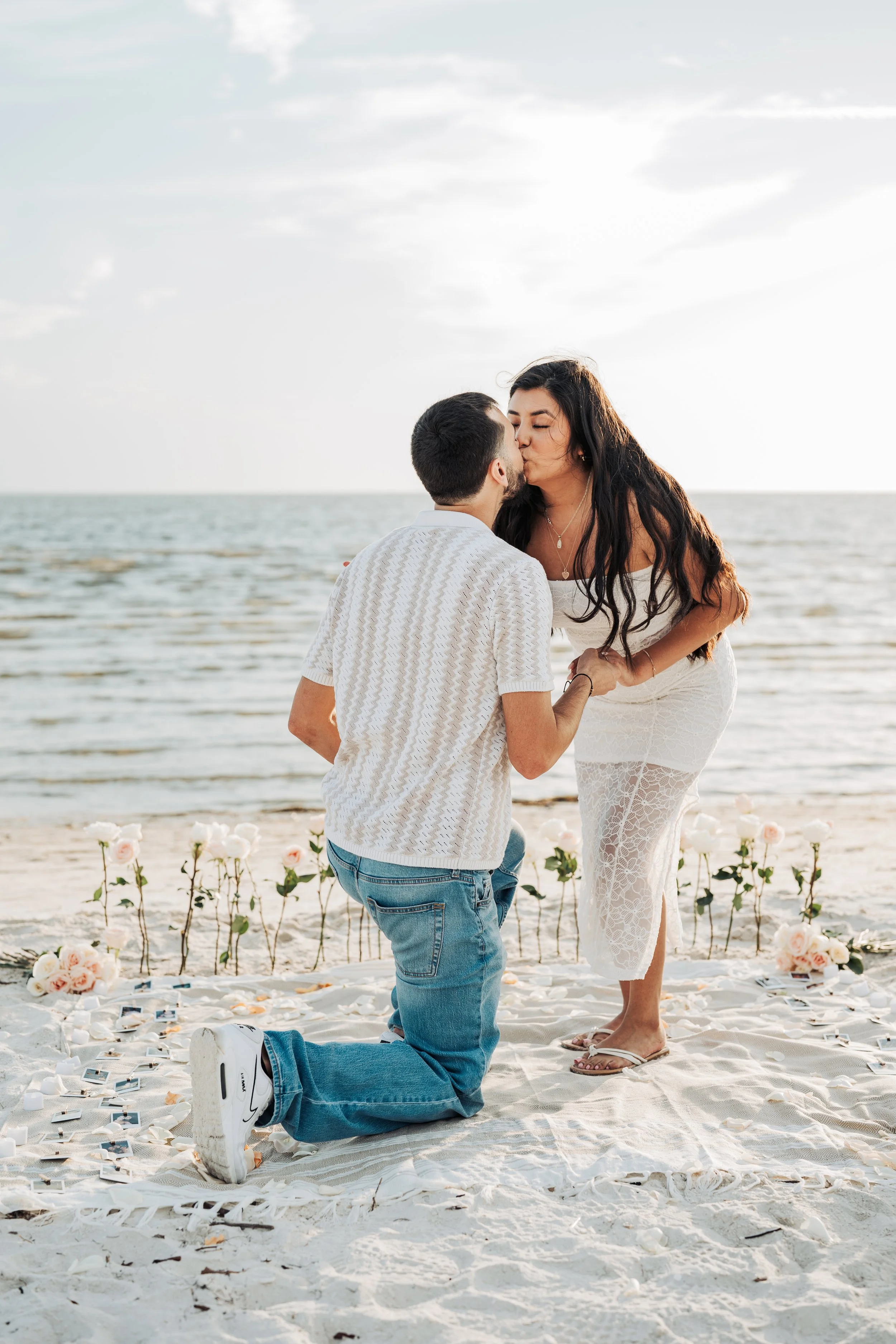 A couple on a beach during sunset, with the man kneeling and proposing marriage to the woman, surrounded by flower petals and photos.