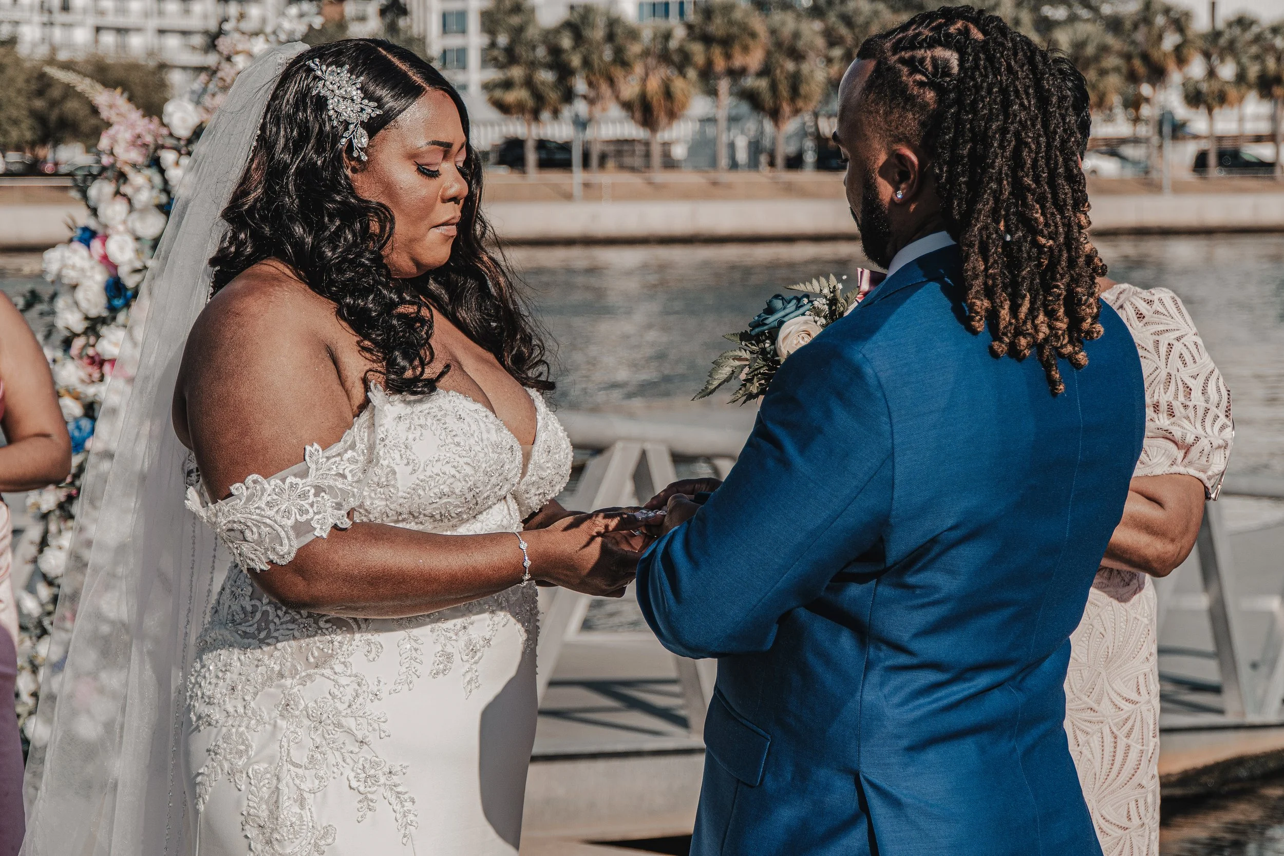 A bride and groom exchange wedding vows outdoors near a river, with a floral arch behind them and a cityscape in the background.
