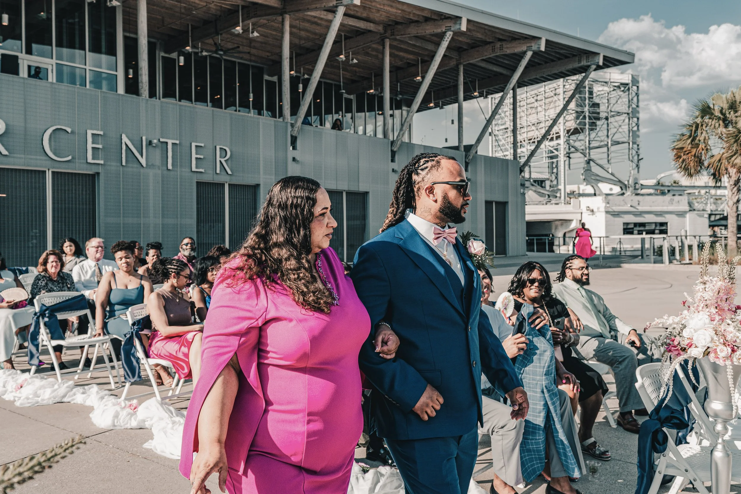 A diverse group of people attending an outdoor ceremony, with some standing and others seated, in front of a modern building with large windows and palm trees.