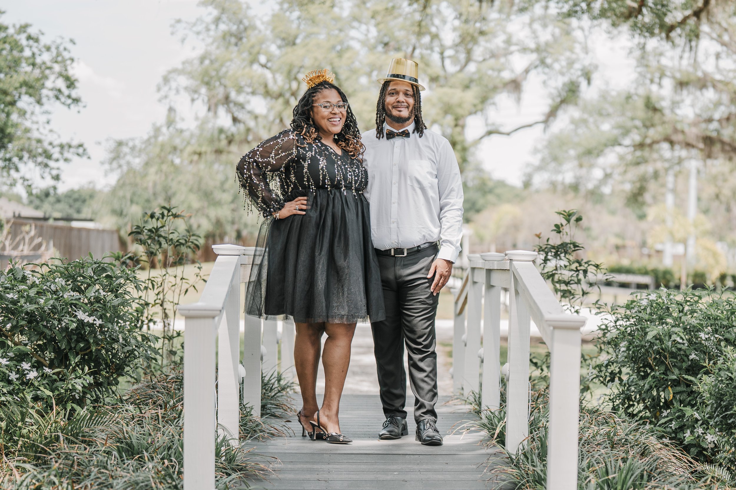 A smiling woman and man standing on a wooden bridge outdoors, dressed in festive attire, with the woman wearing a 'Happy New Year' headband and the man wearing a gold hat and bow tie, surrounded by greenery and trees.