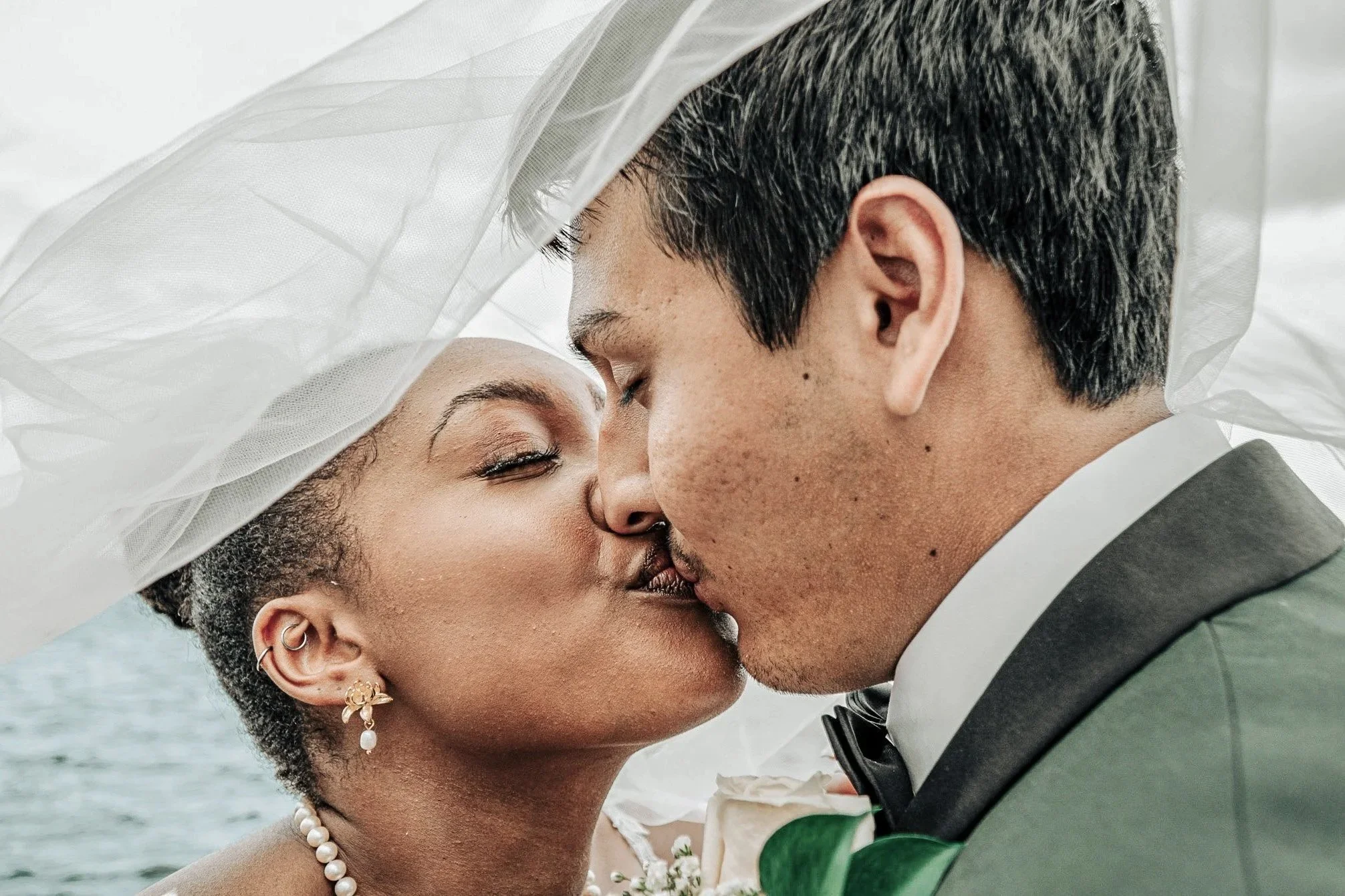 Close-up of a married couple about to kiss, with a white veil over the woman and an outdoor water background.