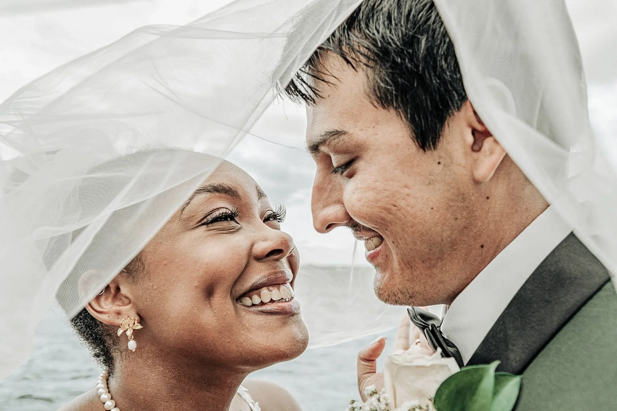 A close-up of a smiling bride and groom with their foreheads touching, under a white veil, during their wedding.