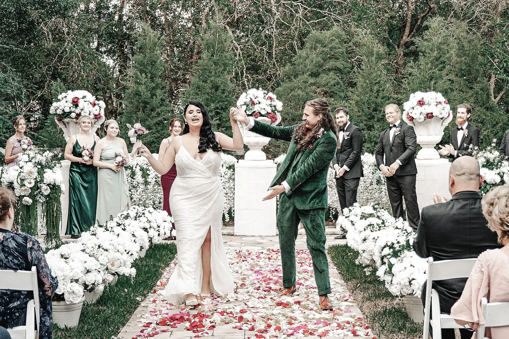 A wedding ceremony outdoors with a bride and groom holding hands at the altar, surrounded by bridesmaids and groomsmen, decorated with large floral arrangements and a flower-strewn aisle.