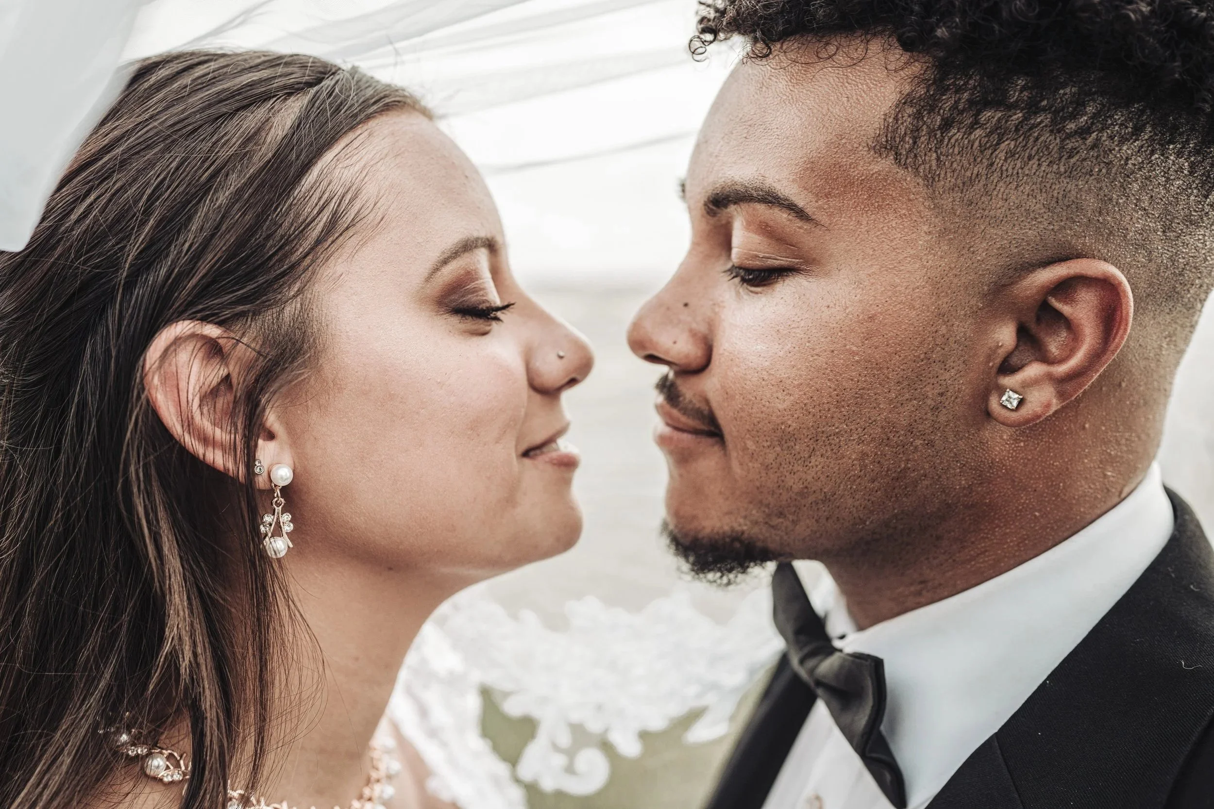 Close-up of a couple touching foreheads, eyes closed, smiling, with wedding attire, outdoors.