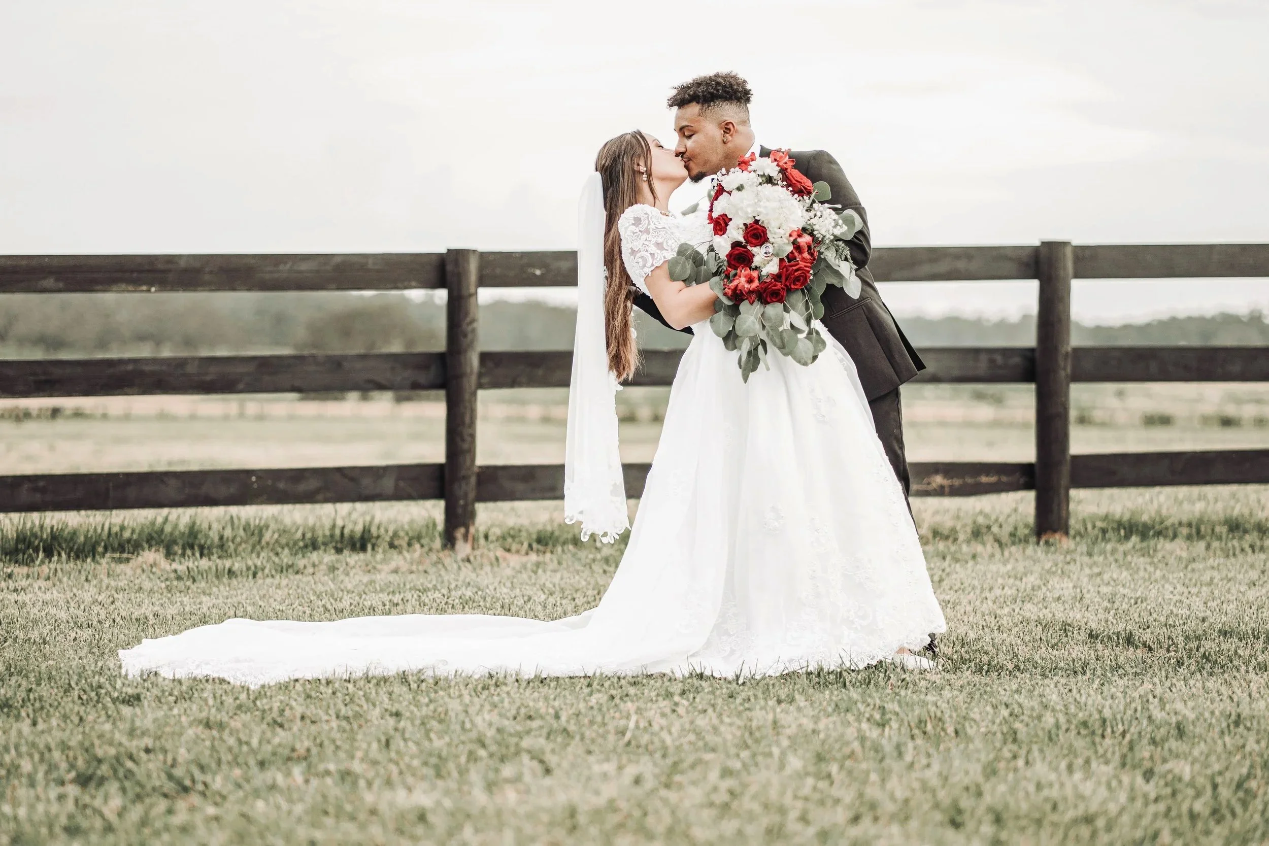 A bride and groom sharing a kiss outdoors on their wedding day, with the bride holding a large bouquet of red and white flowers, and a wooden fence in the background.