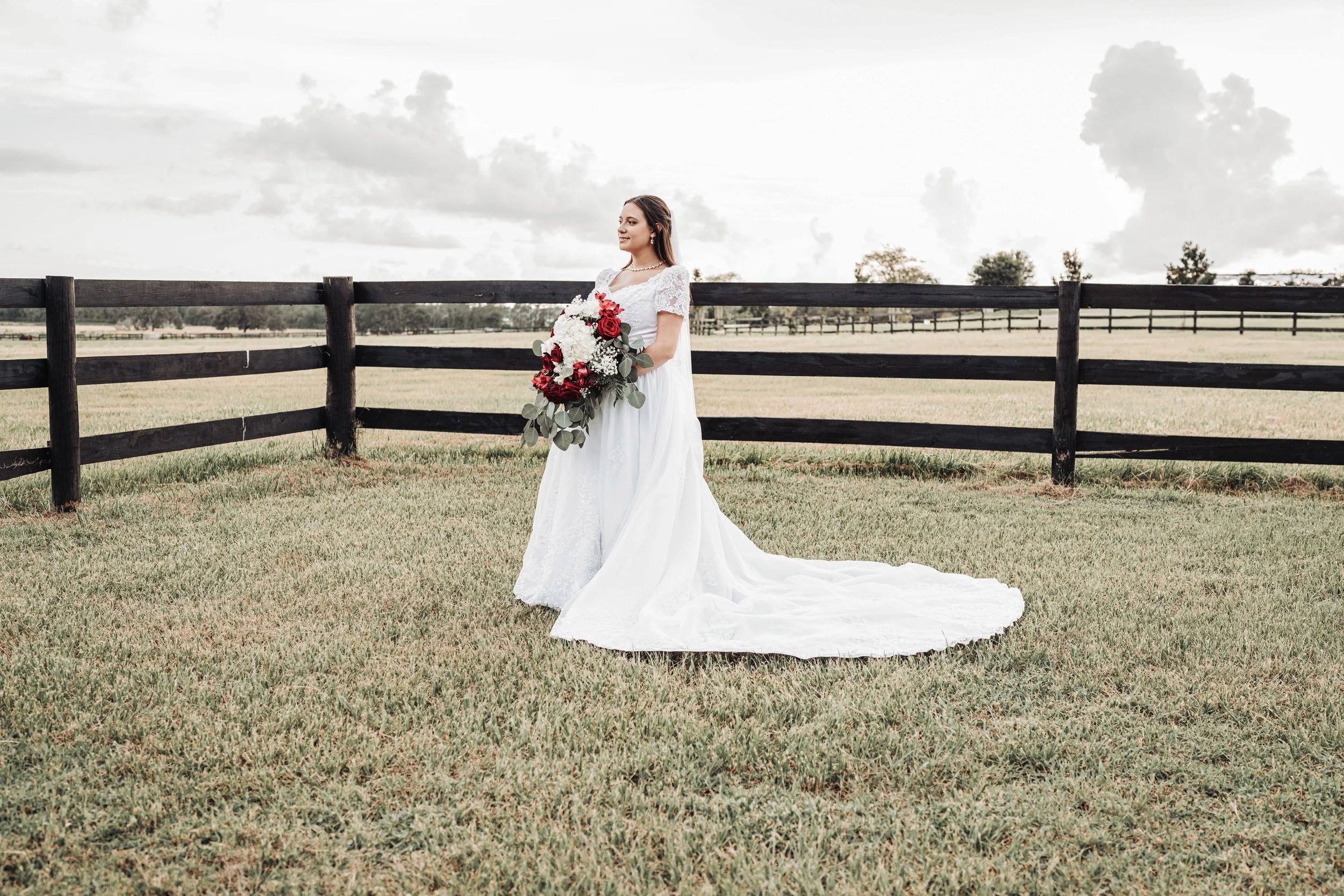 A bride in a white wedding dress holding a bouquet of red and white flowers standing outdoors by a black wooden fence, with a cloudy sky and trees in the background.