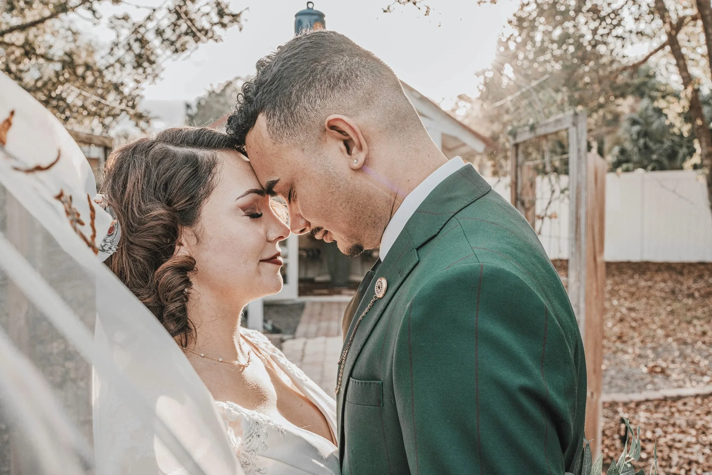 A couple stands closely with foreheads touching, eyes closed, outdoors on a sunny day during a wedding or romantic event. The woman has dark, curly hair and wears a white dress, while the man has short hair and is dressed in a green suit. She is hold